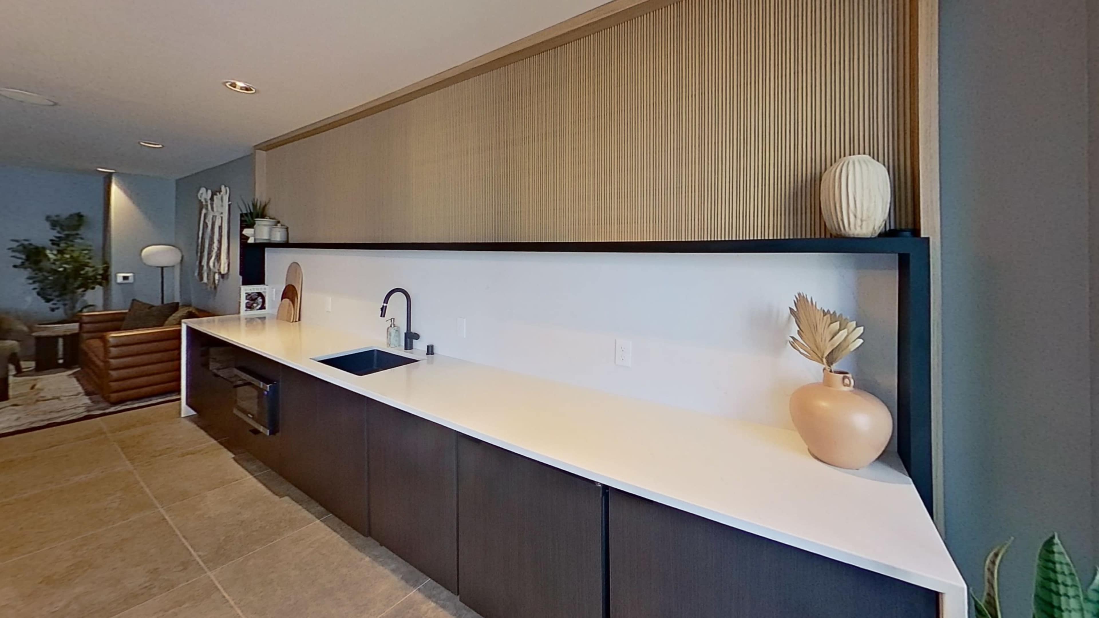 The image shows a modern kitchen with a long countertop, a black sink, and wooden wall panels, complemented by decorative vases.