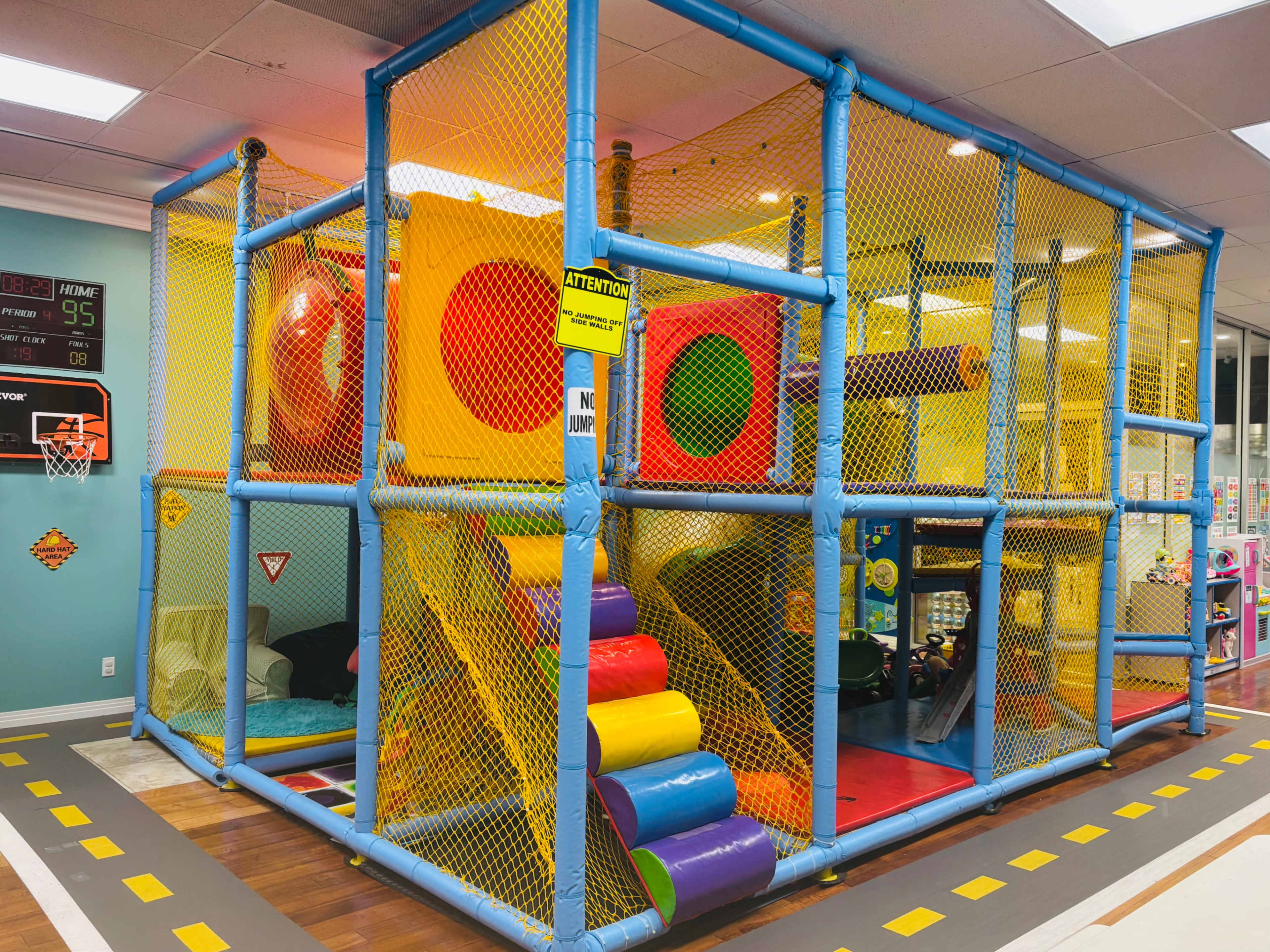 The image shows an indoor play structure enclosed by a yellow net, featuring colorful slides, tunnels, and various climbing areas.