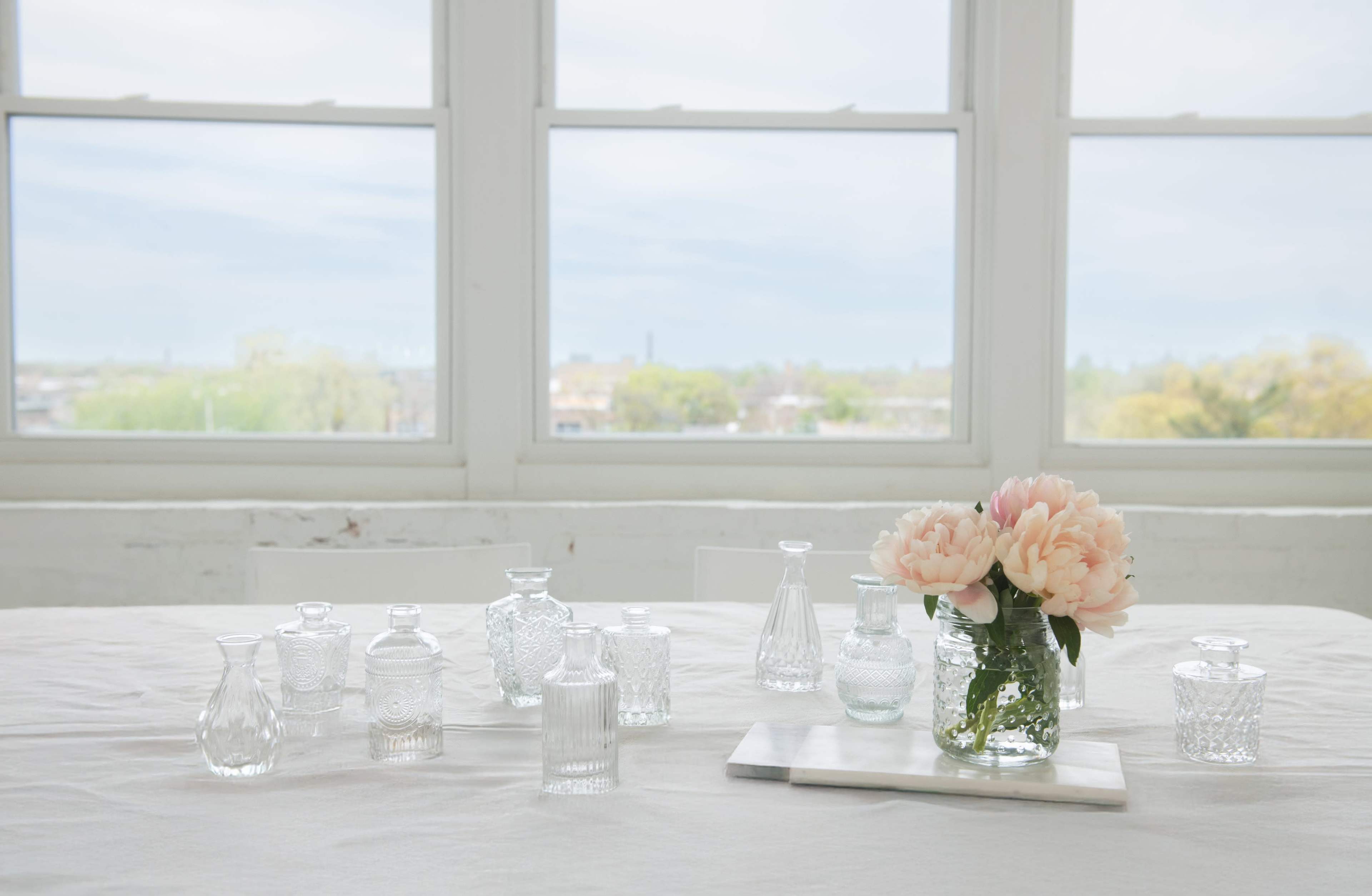 A collection of glass vases sits on a light-colored table, with a bouquet of pink peonies in a jar, while large windows frame a view of greenery outside.