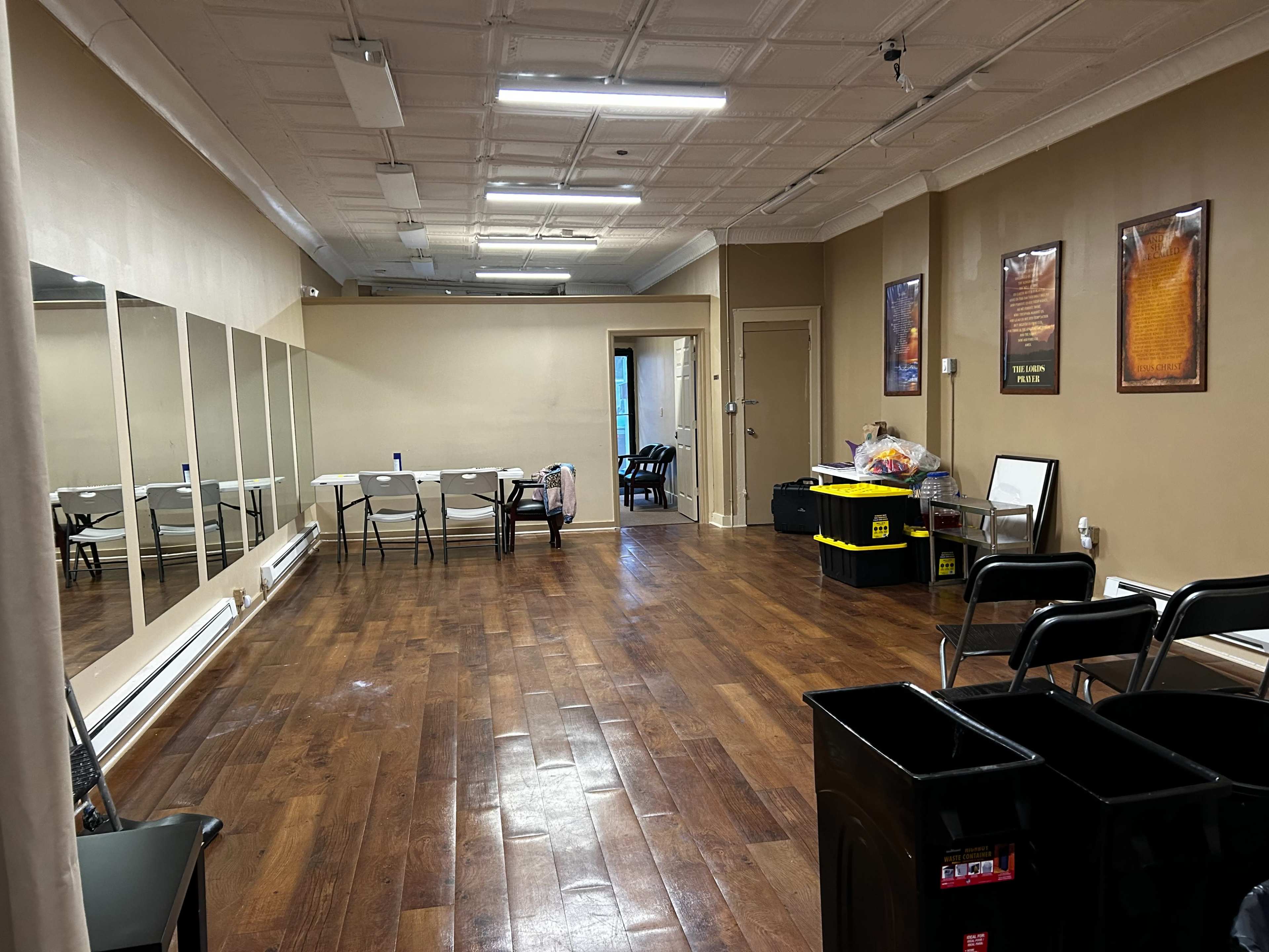 An empty room with wooden flooring, a large mirror on one wall, and several folding chairs arranged along the side.