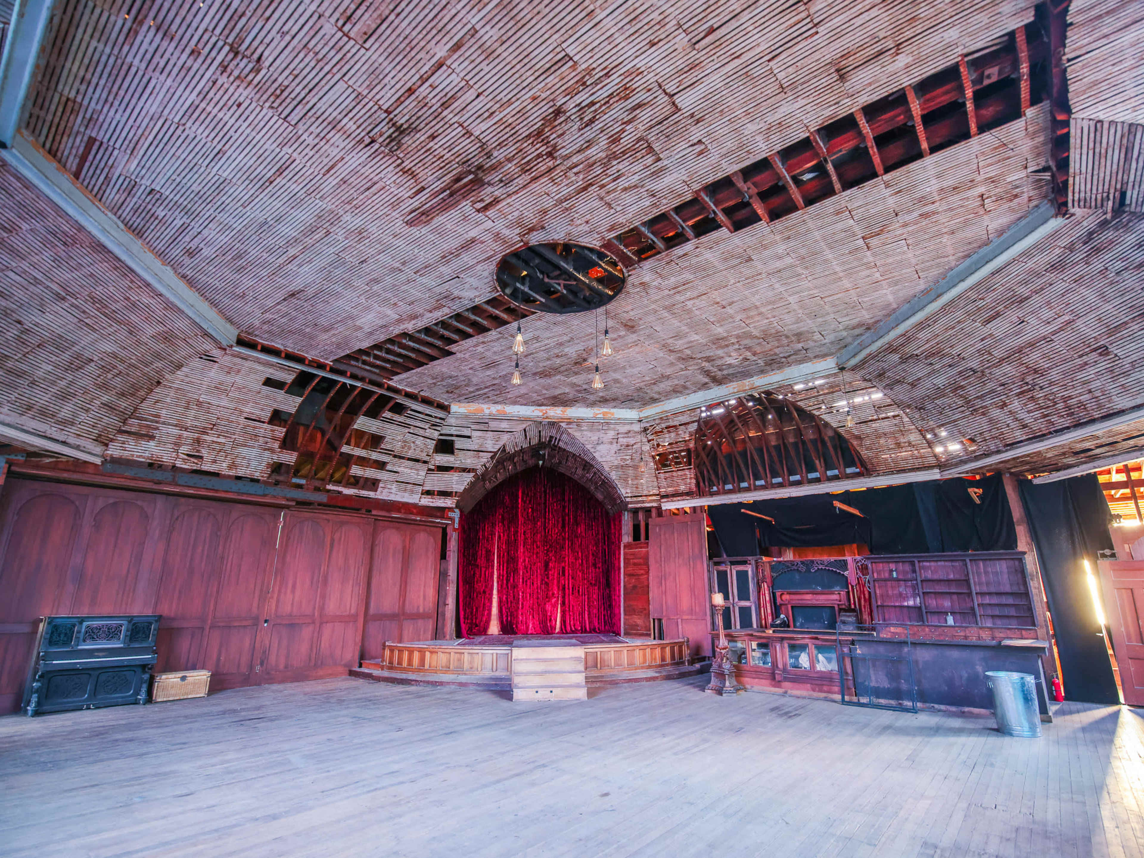 An empty stage with a red velvet curtain in a wooden room featuring a high, arched ceiling and exposed beams.