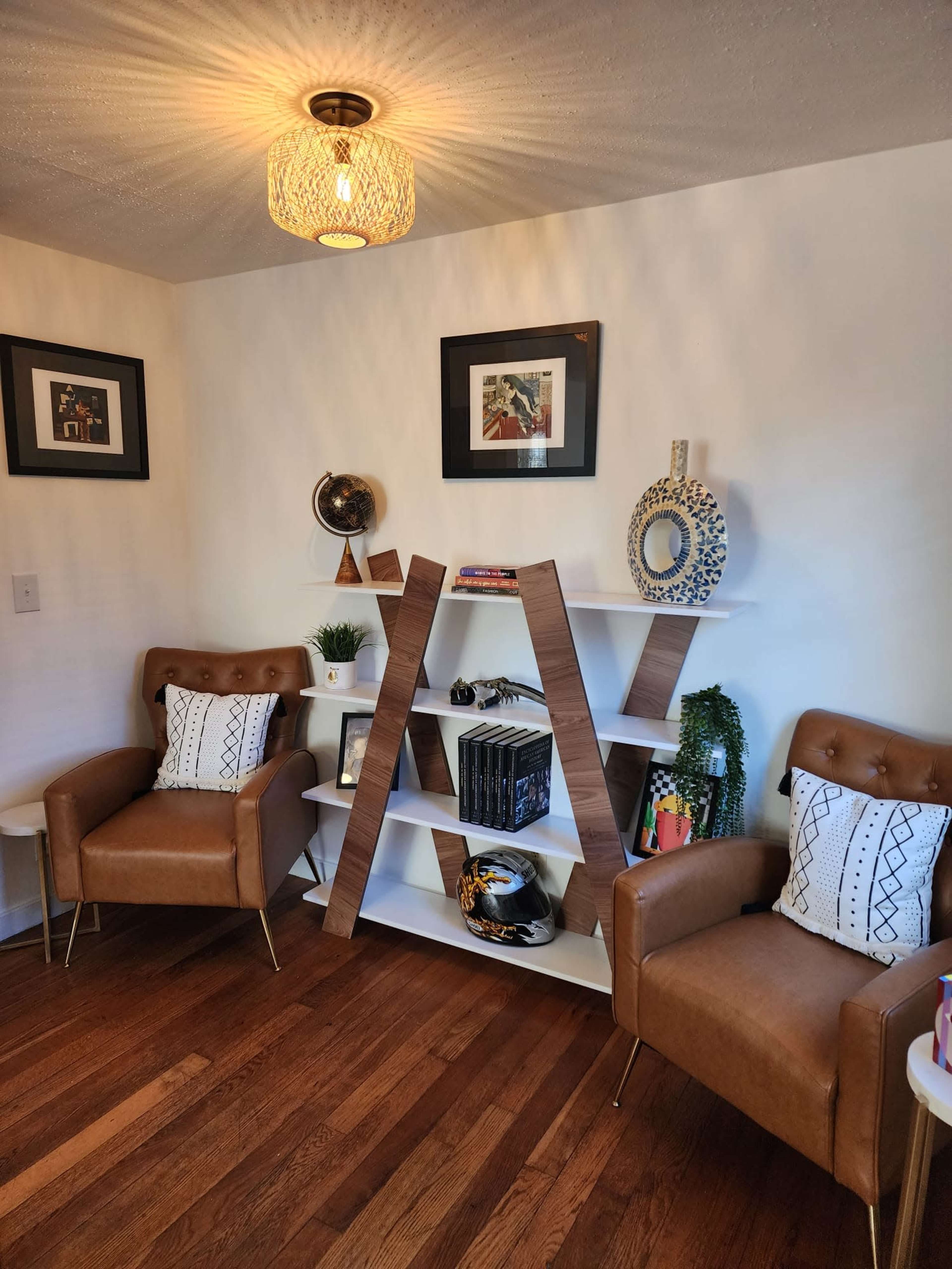 A cozy corner featuring two brown leather armchairs with decorative pillows, a leaning bookshelf adorned with books and ornaments, and a light fixture hanging from the ceiling.