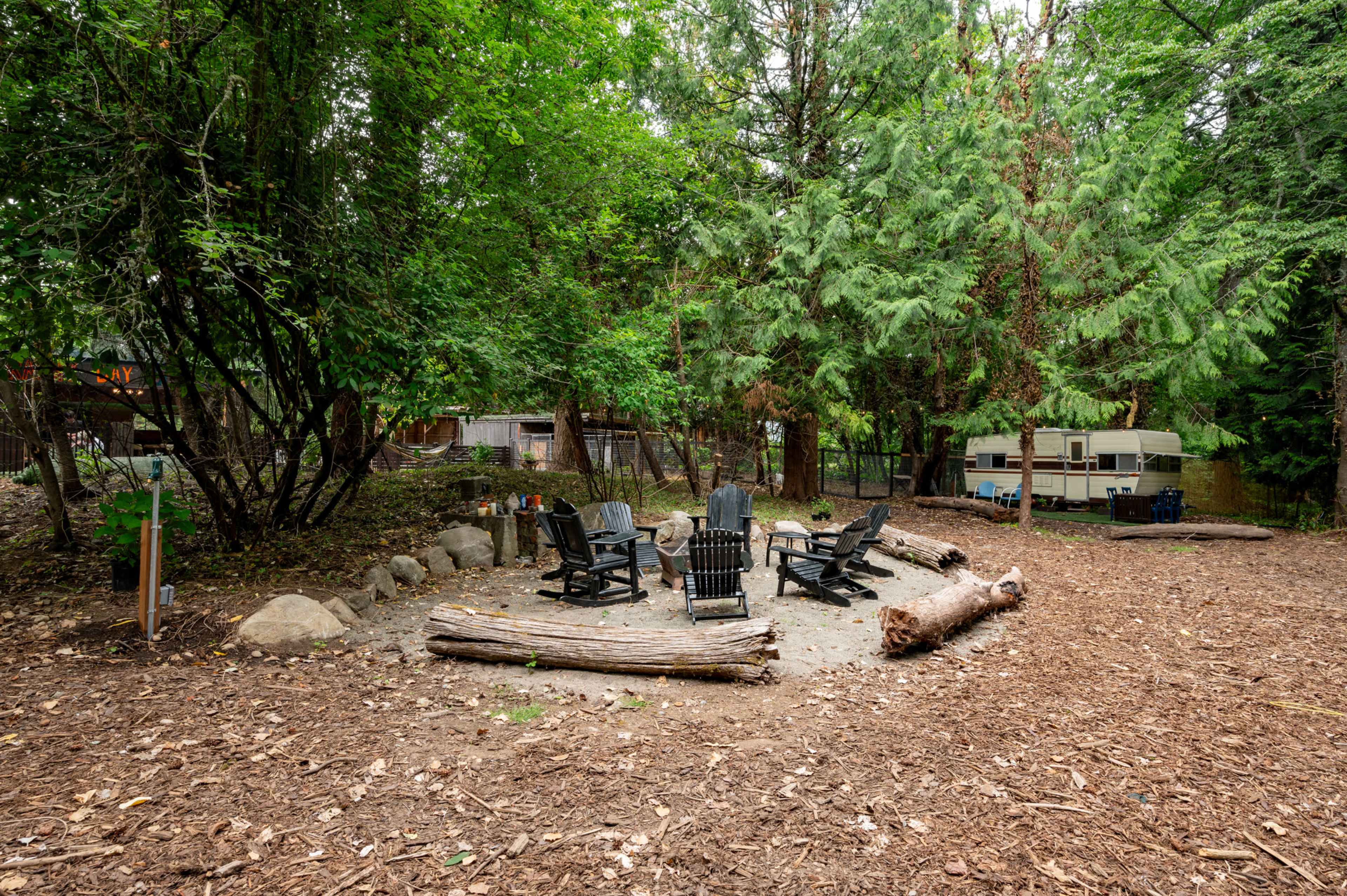 A campfire circle with several black chairs surrounds a sandy area in a wooded setting.