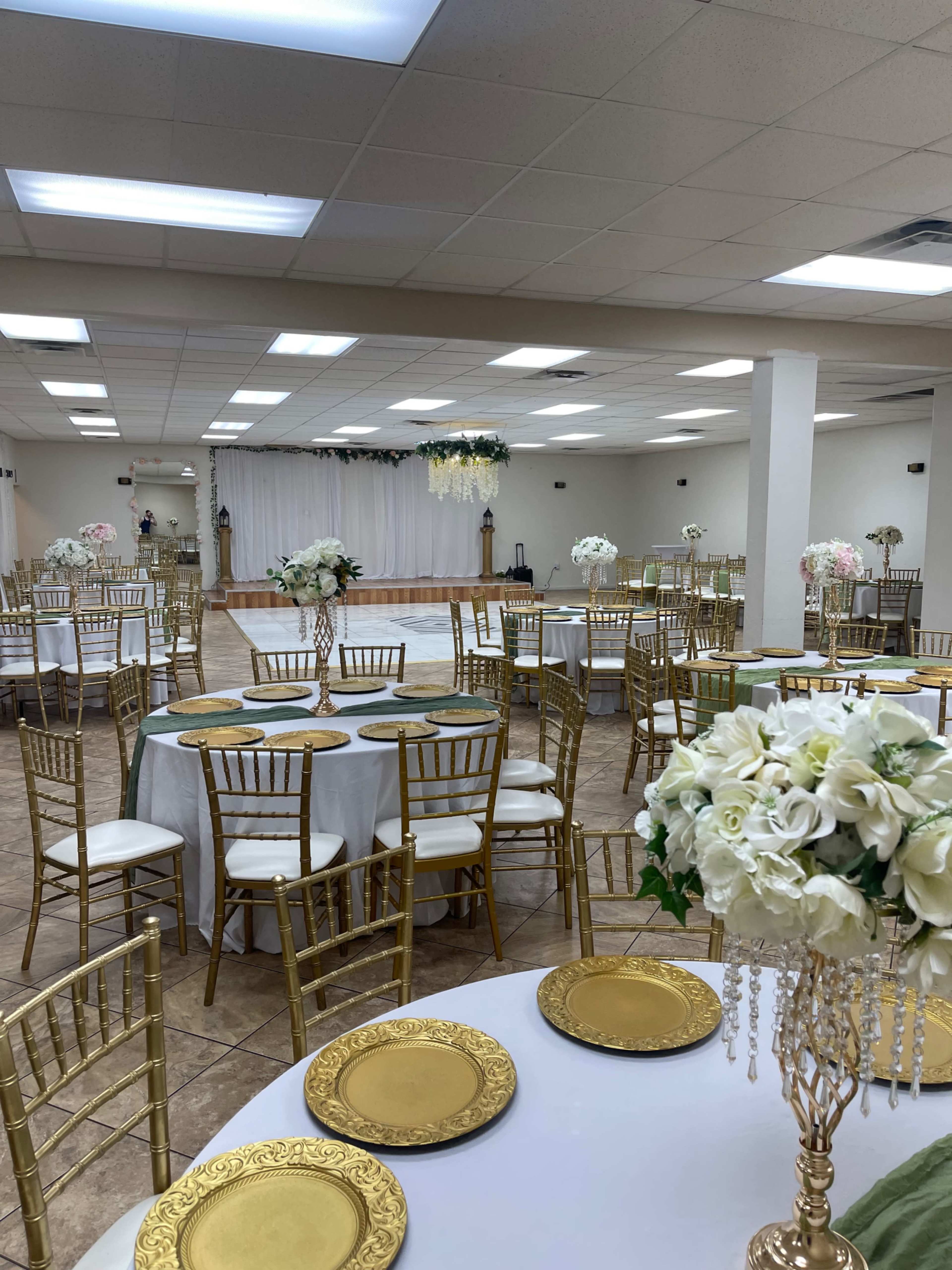 The image shows a banquet hall set up for an event, featuring round tables with white tablecloths, gold plate settings, and floral centerpieces, with a decorated stage in the background.
