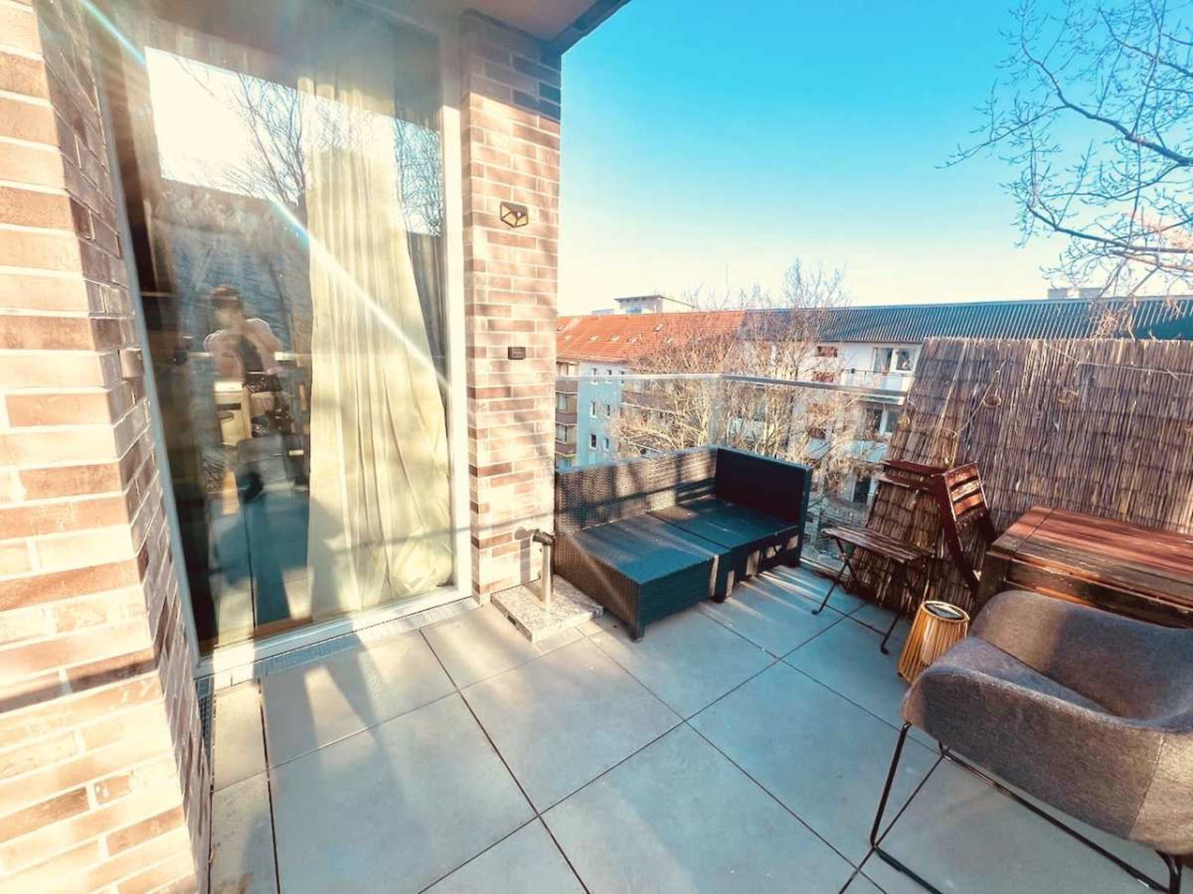 The image shows a balcony with a combination of seating, including a black composite sofa, a wooden chair, and a small side table, surrounded by a view of nearby buildings and trees.