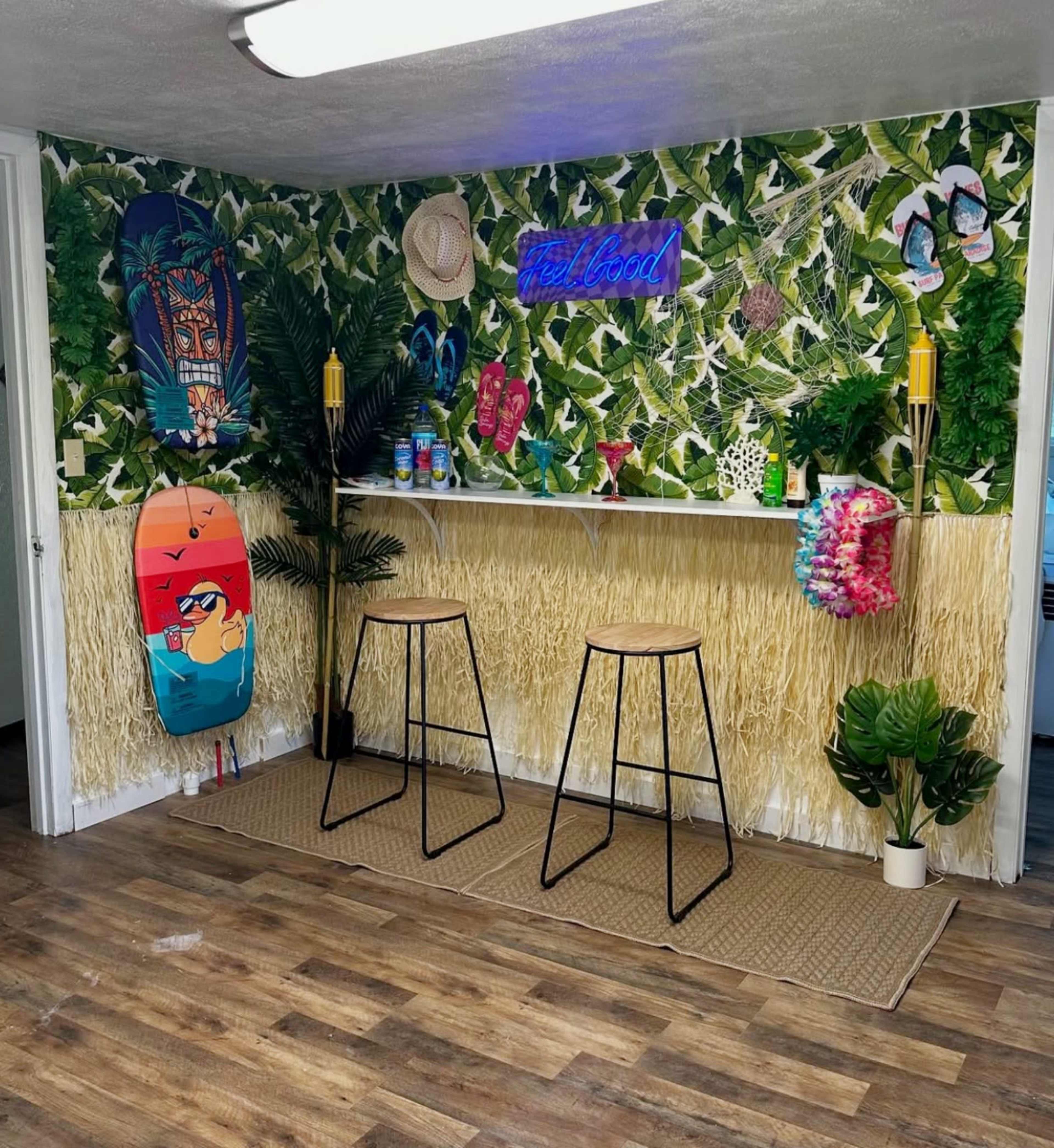 The image shows a tropical-themed corner with two bar stools, a shelf decorated with plants and colorful beach items, and a backdrop of green foliage wallpaper.