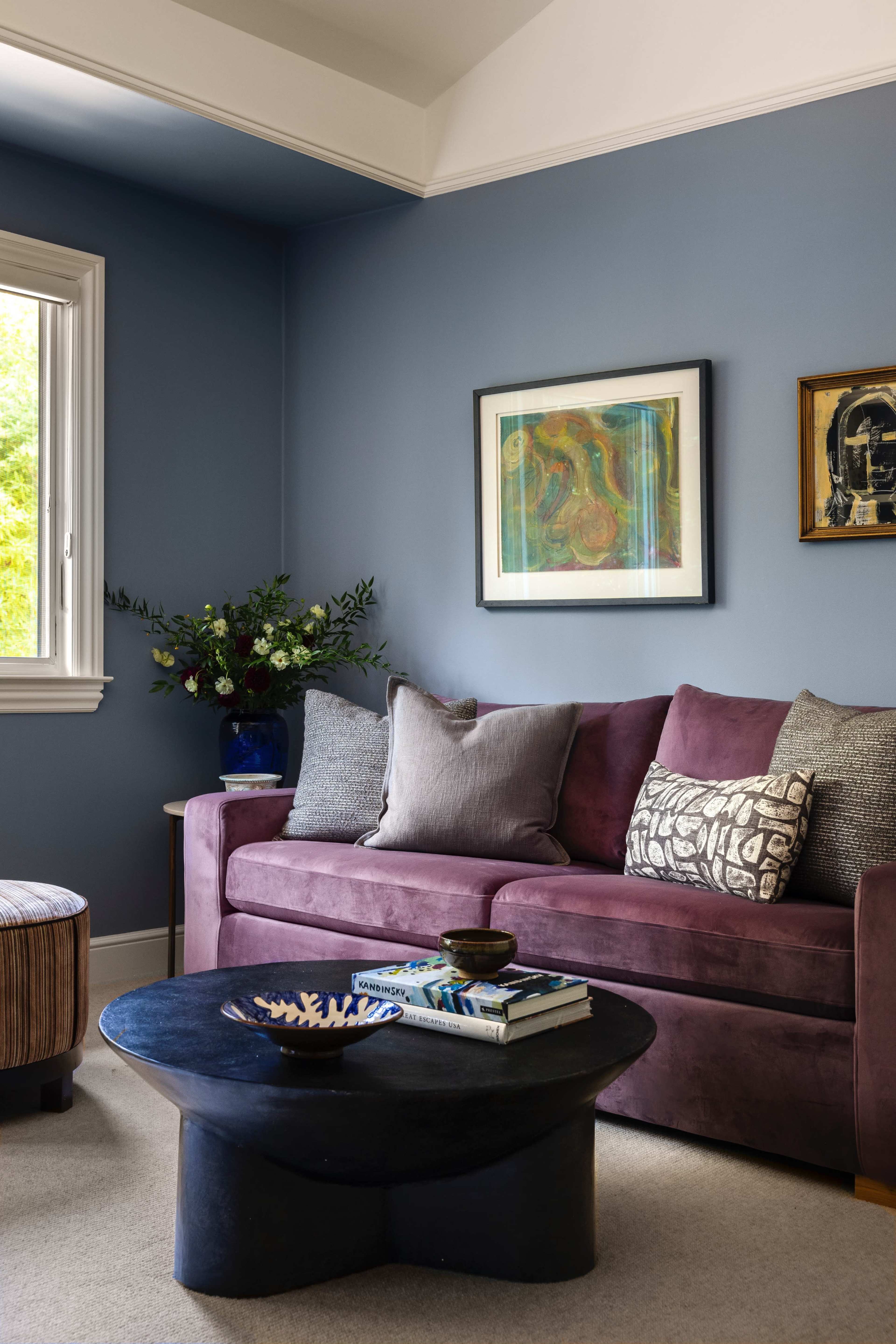 A purple sofa with patterned cushions is set against a blue wall, accompanied by a round black coffee table and a large window with a floral arrangement nearby.