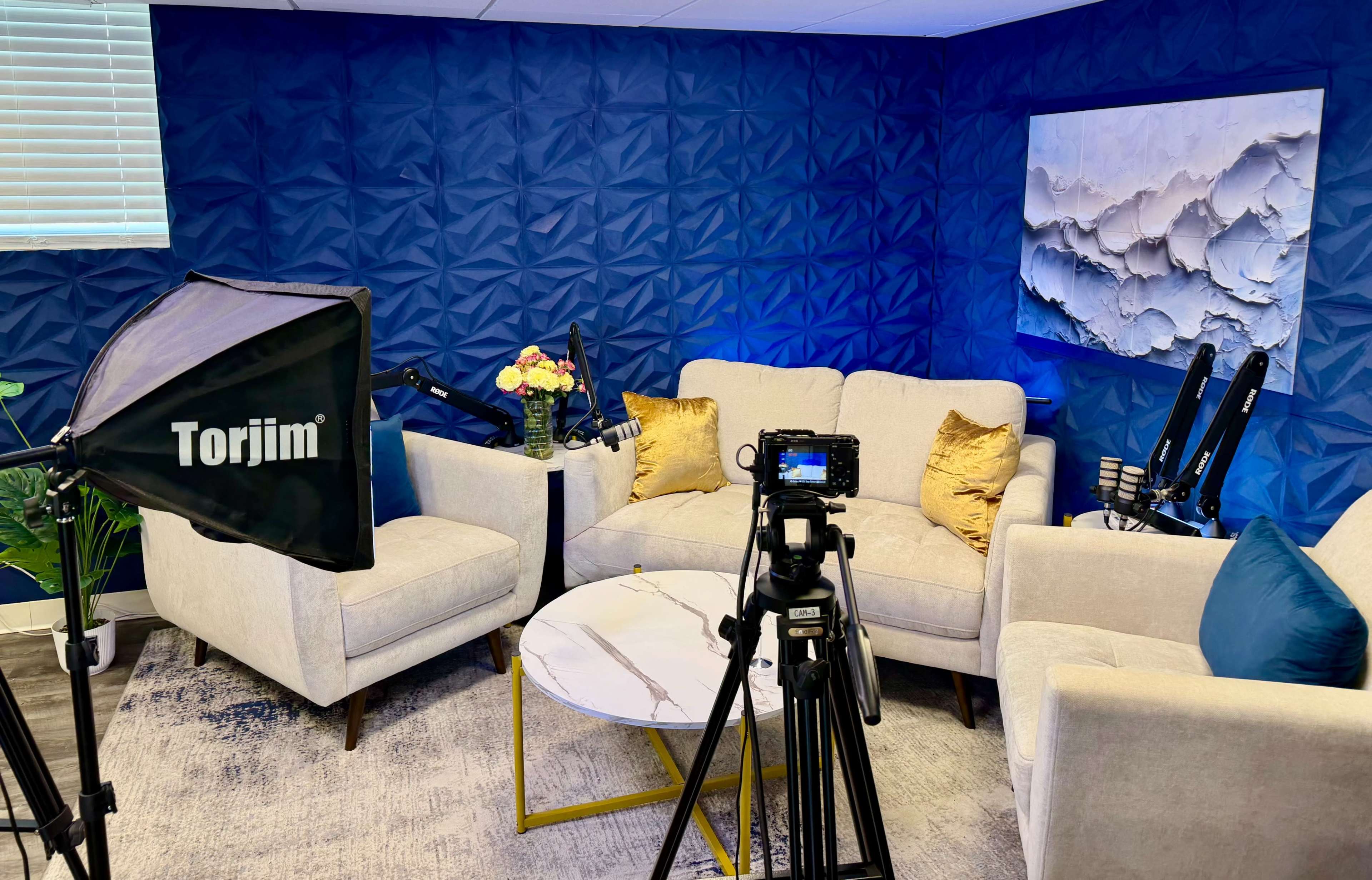 A well-lit recording studio features a blue textured wall, two cream-colored sofas with decorative pillows, a round marble coffee table, and camera equipment set up for filming.
