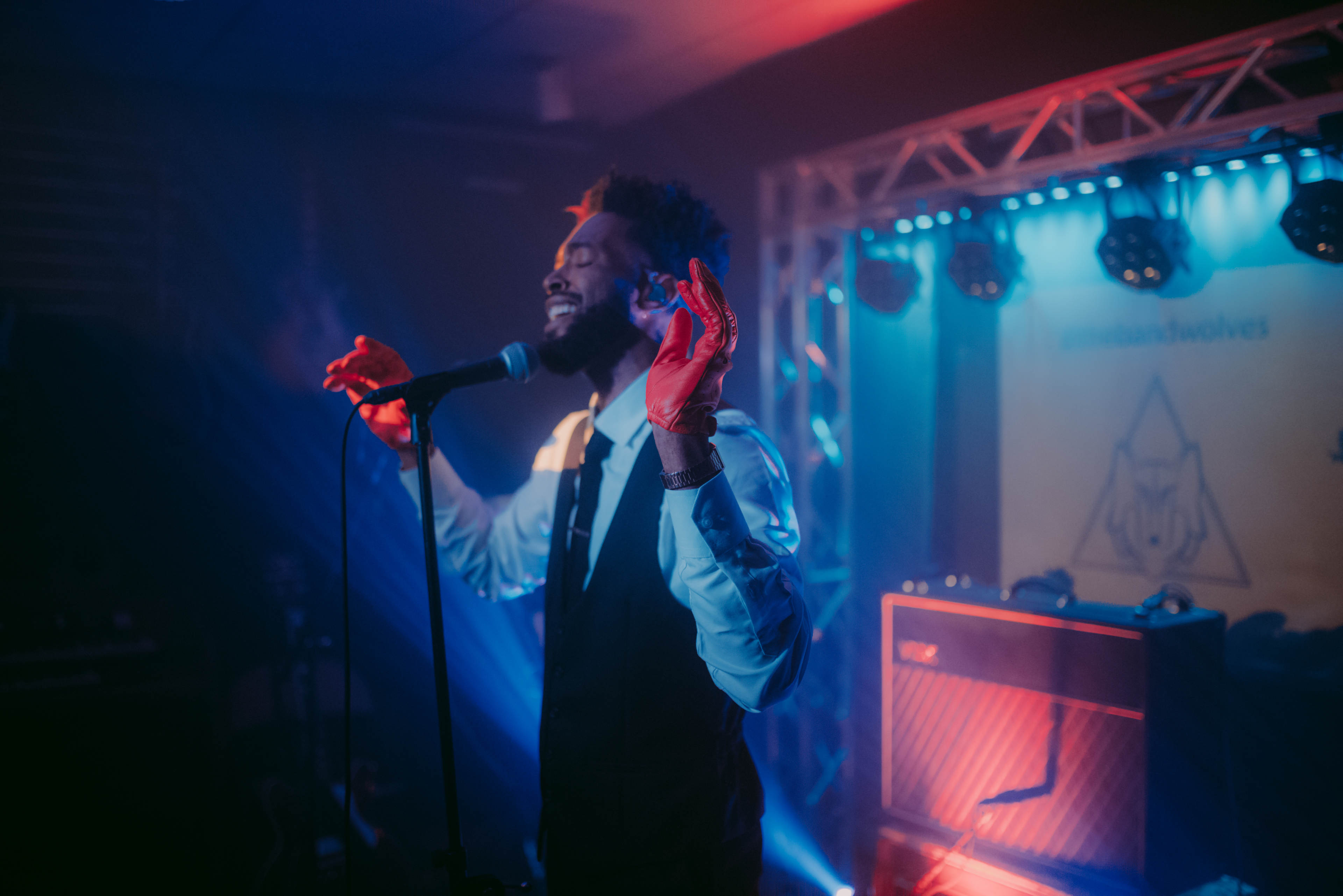 A man wearing a suit sings into a microphone on stage with colorful lighting and a guitar amplifier in the background.