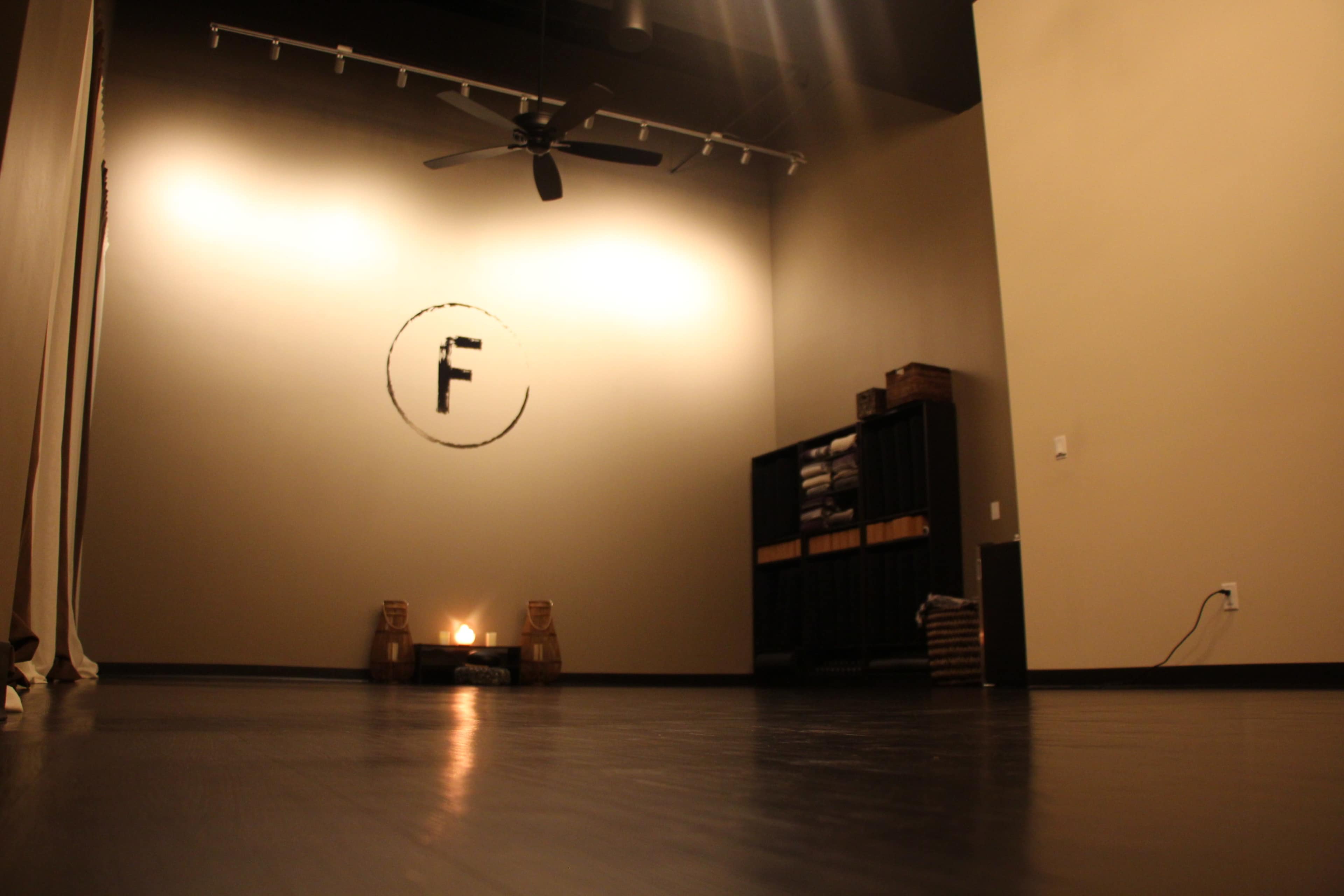 An empty room with a light-colored wall featuring a large letter "F" and a fan hanging from the ceiling, with a shelf of folded towels in the corner.