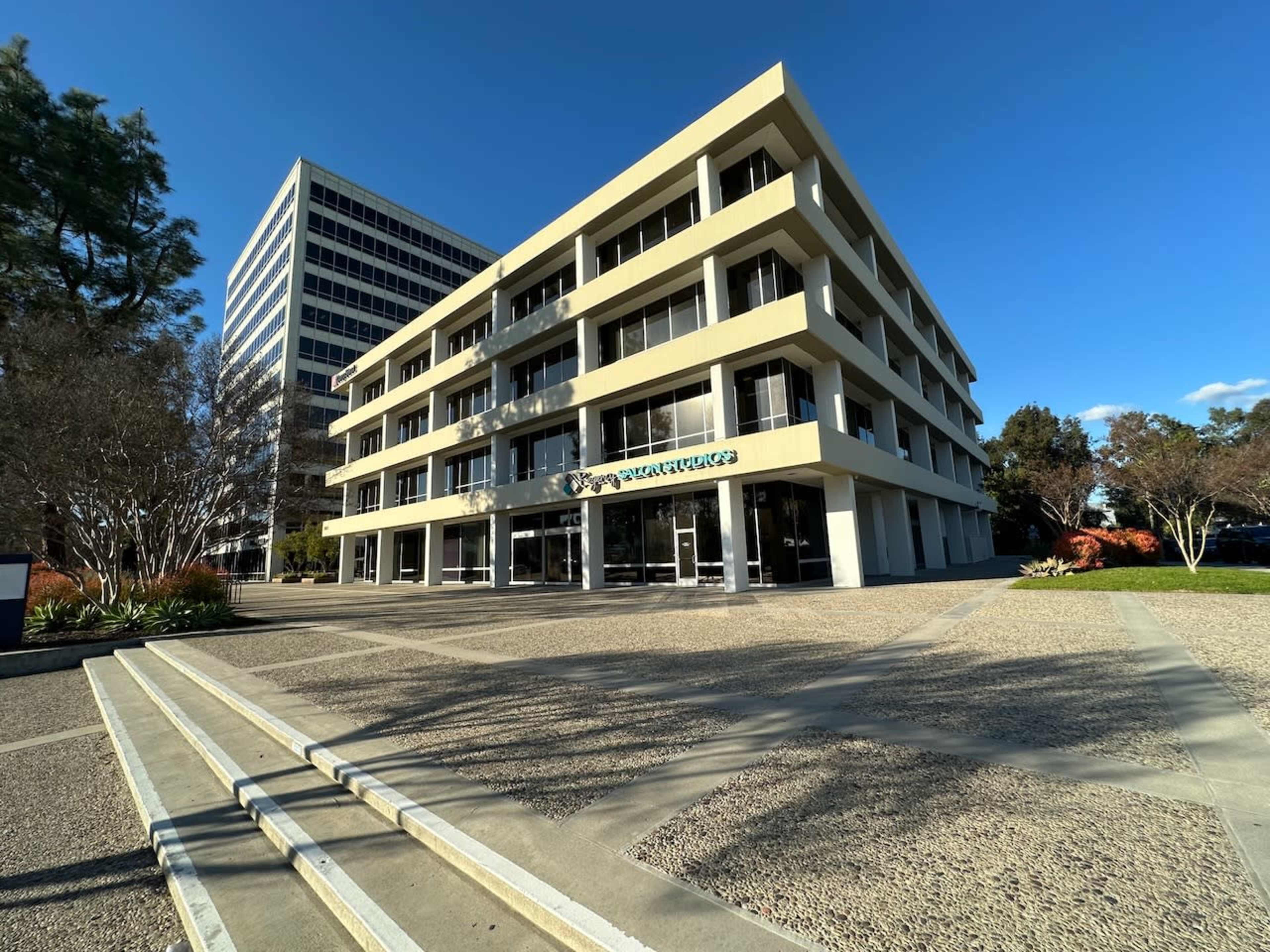 A modern, four-story commercial building with a flat facade and large windows, situated in a landscaped area with a paved walkway.