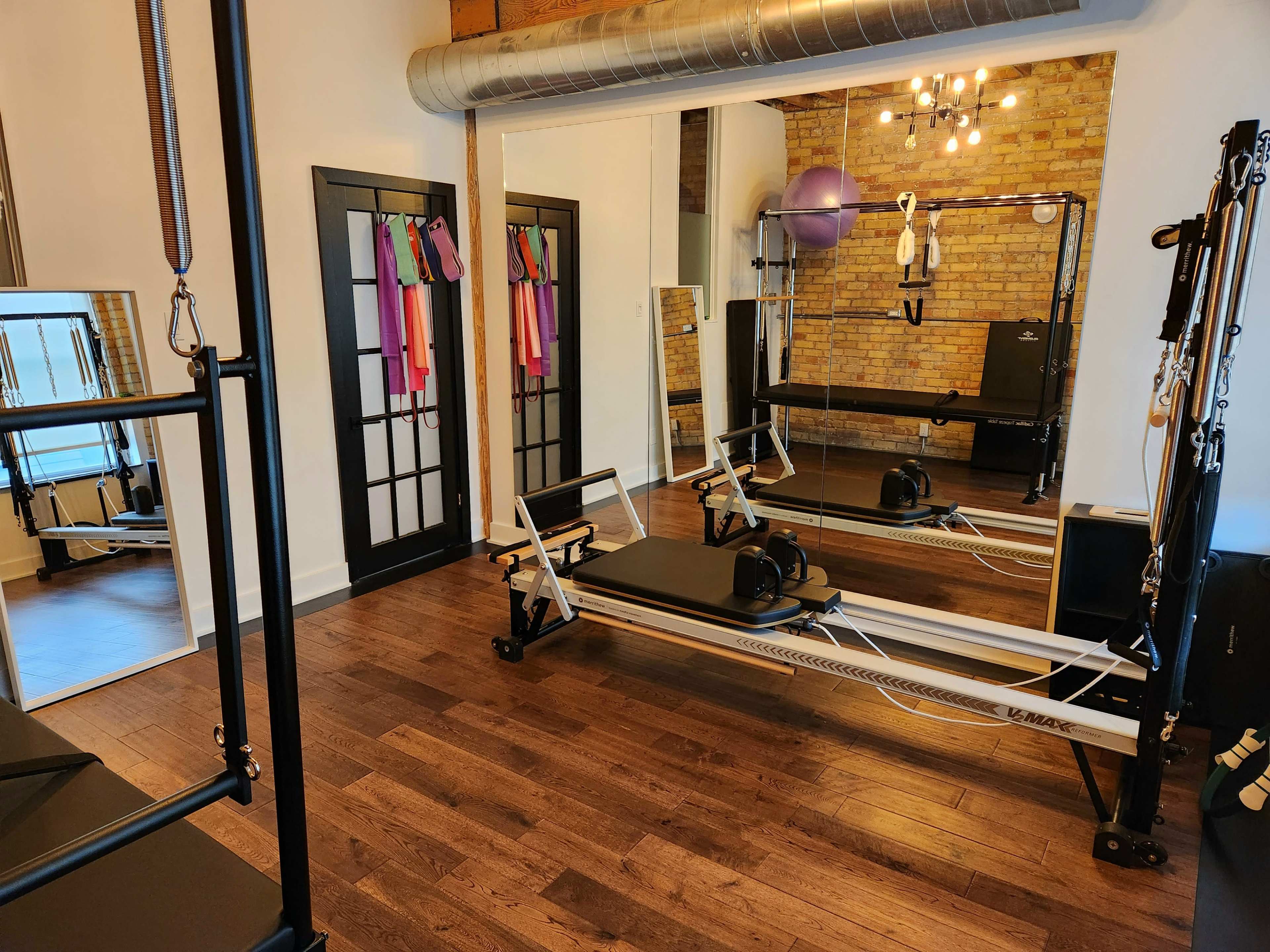 A spacious fitness studio with wooden floors, featuring a reformer machine, mirrors, and exercise equipment against a brick wall.