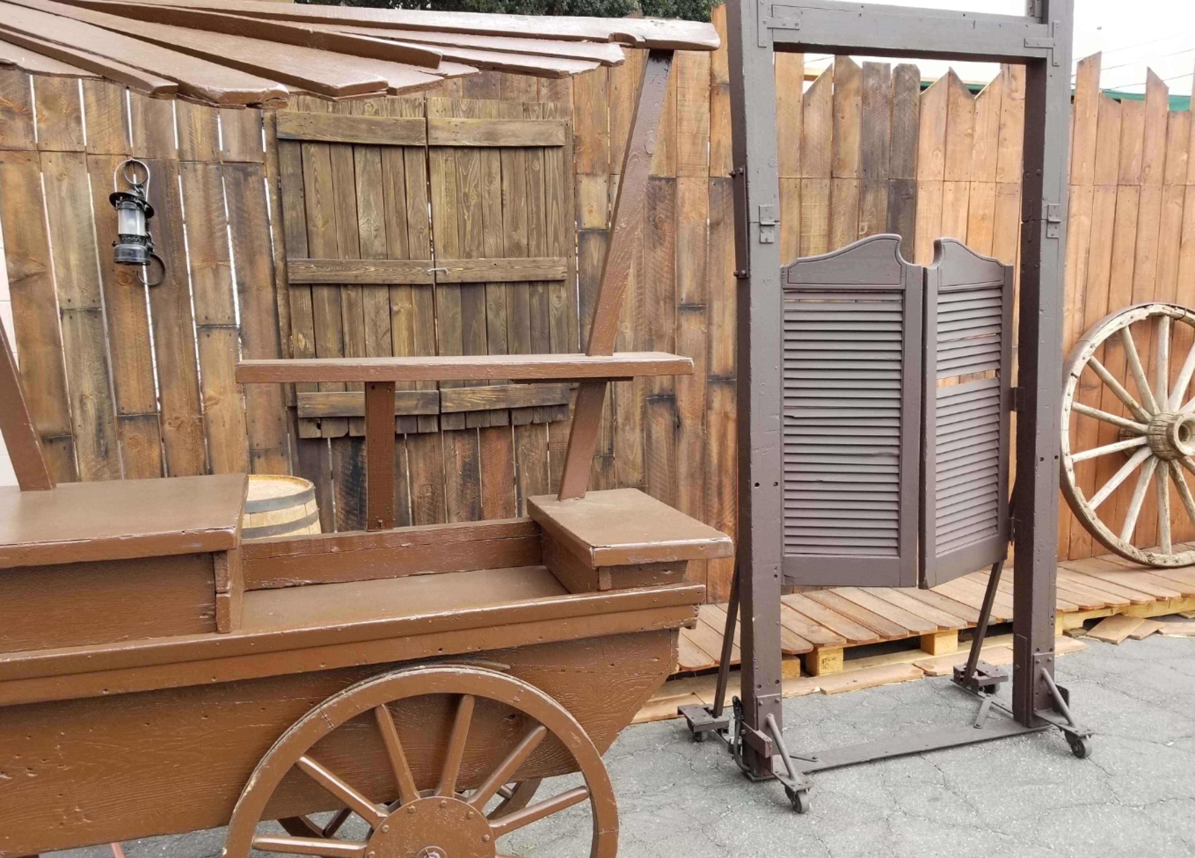 A wooden cart beside a rustic wood fence, with a slatted roof and a wooden gate, along with a wagon wheel in the background.