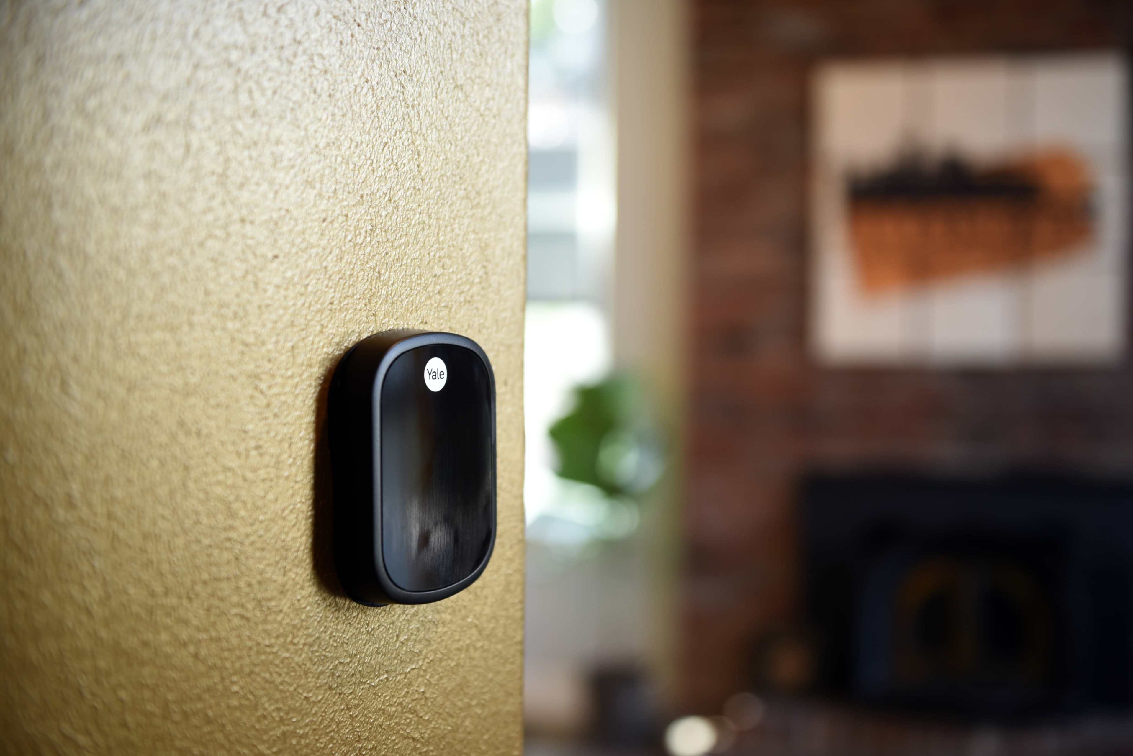 A black smart home device is mounted on a gold wall, with a blurred background featuring a fireplace and artwork.
