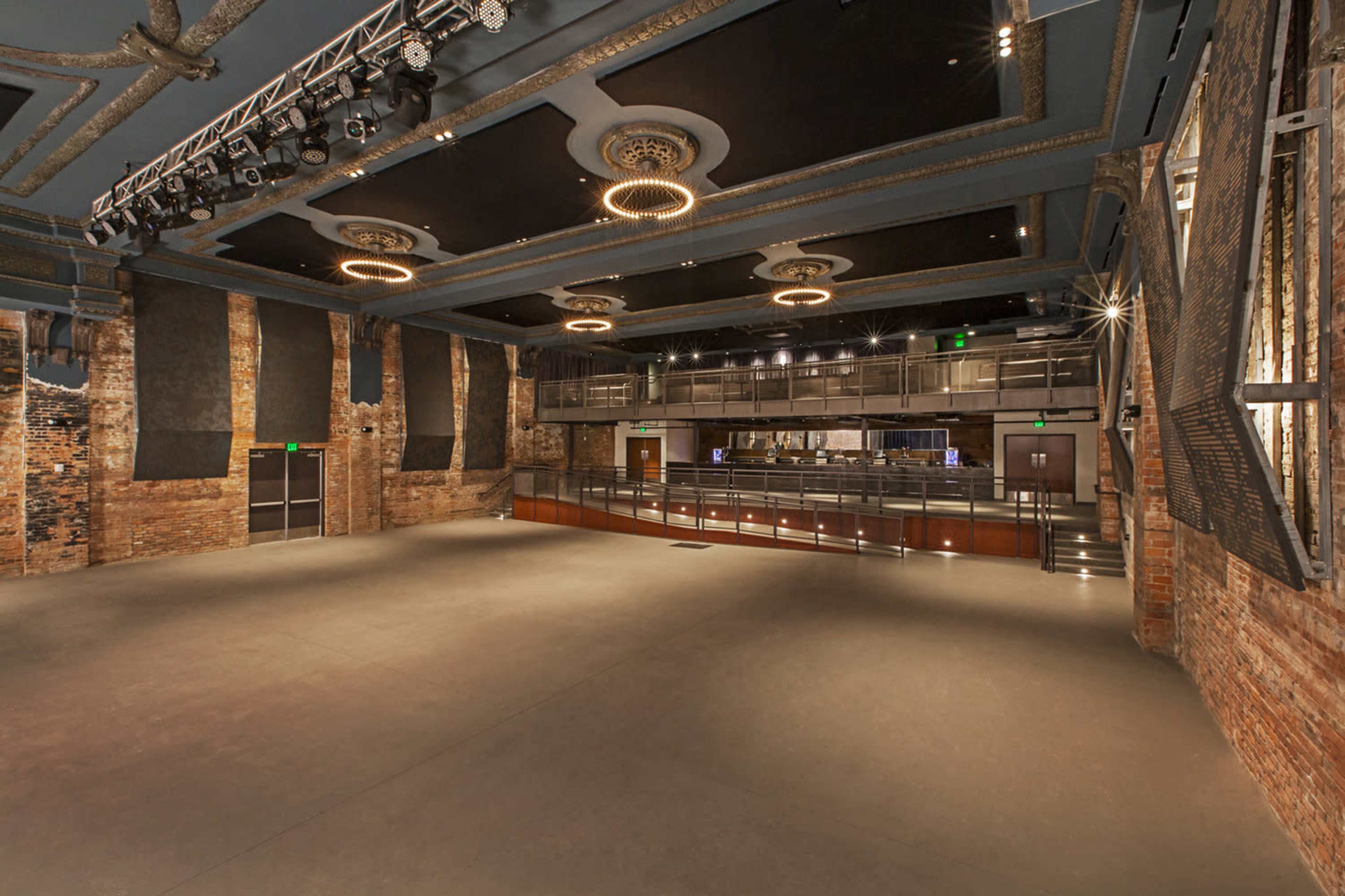 A large, empty concert venue featuring a stage with overhead lighting and a balcony area, with exposed brick walls and a polished floor.