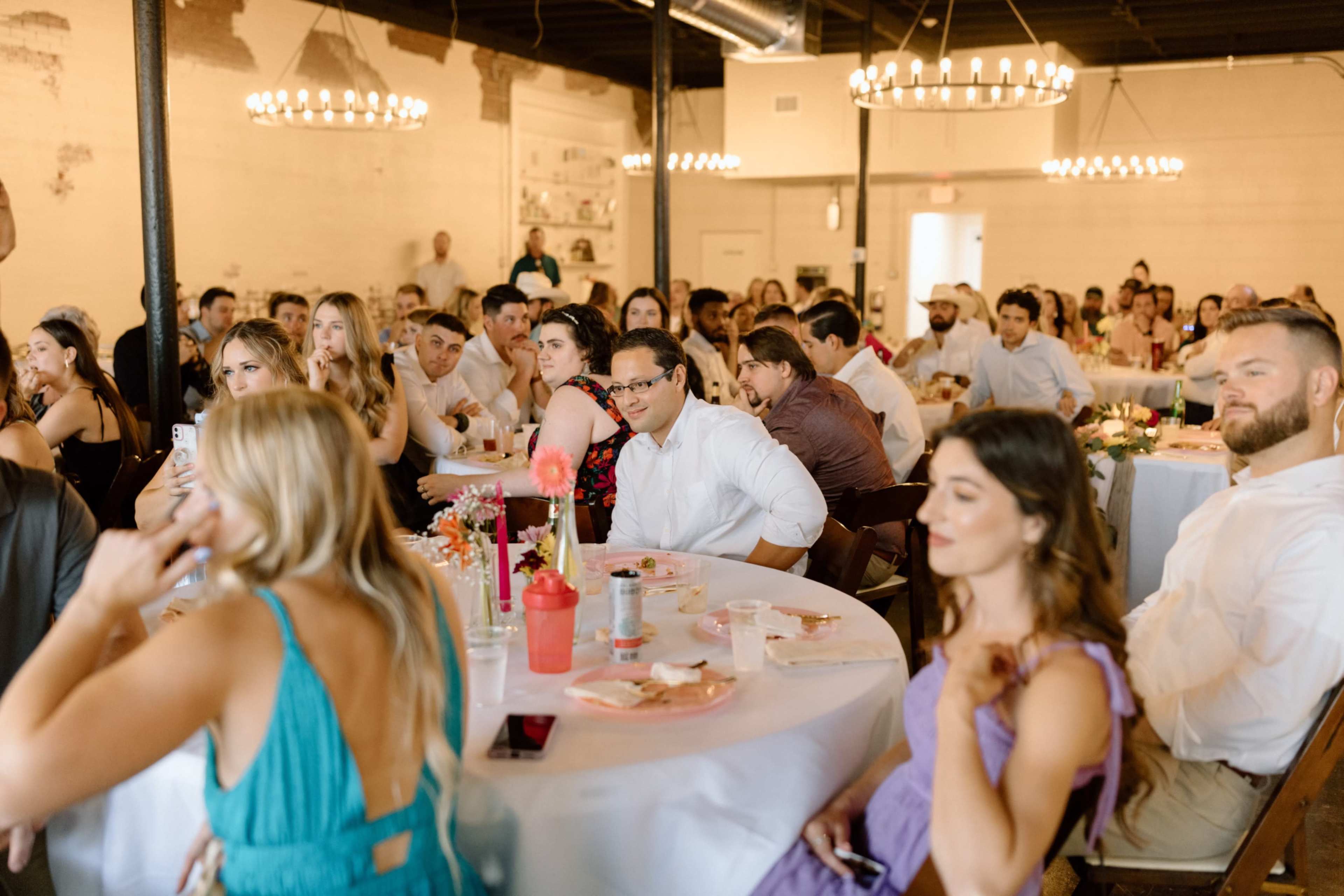 A group of guests sit at elegantly dressed tables during a gathering in a well-lit venue.