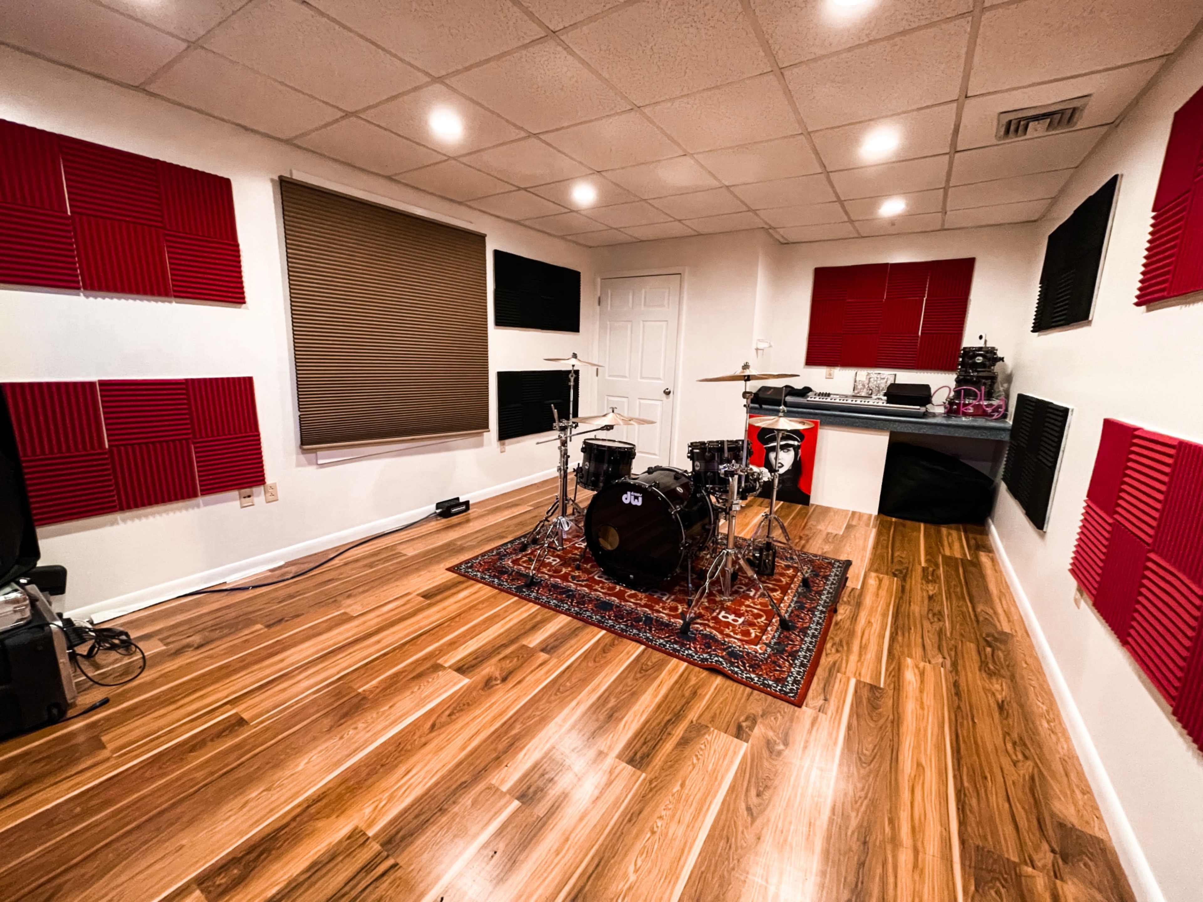The image shows a music rehearsal room with a black drum set on a rug, surrounded by soundproofing panels on the walls and wooden flooring.