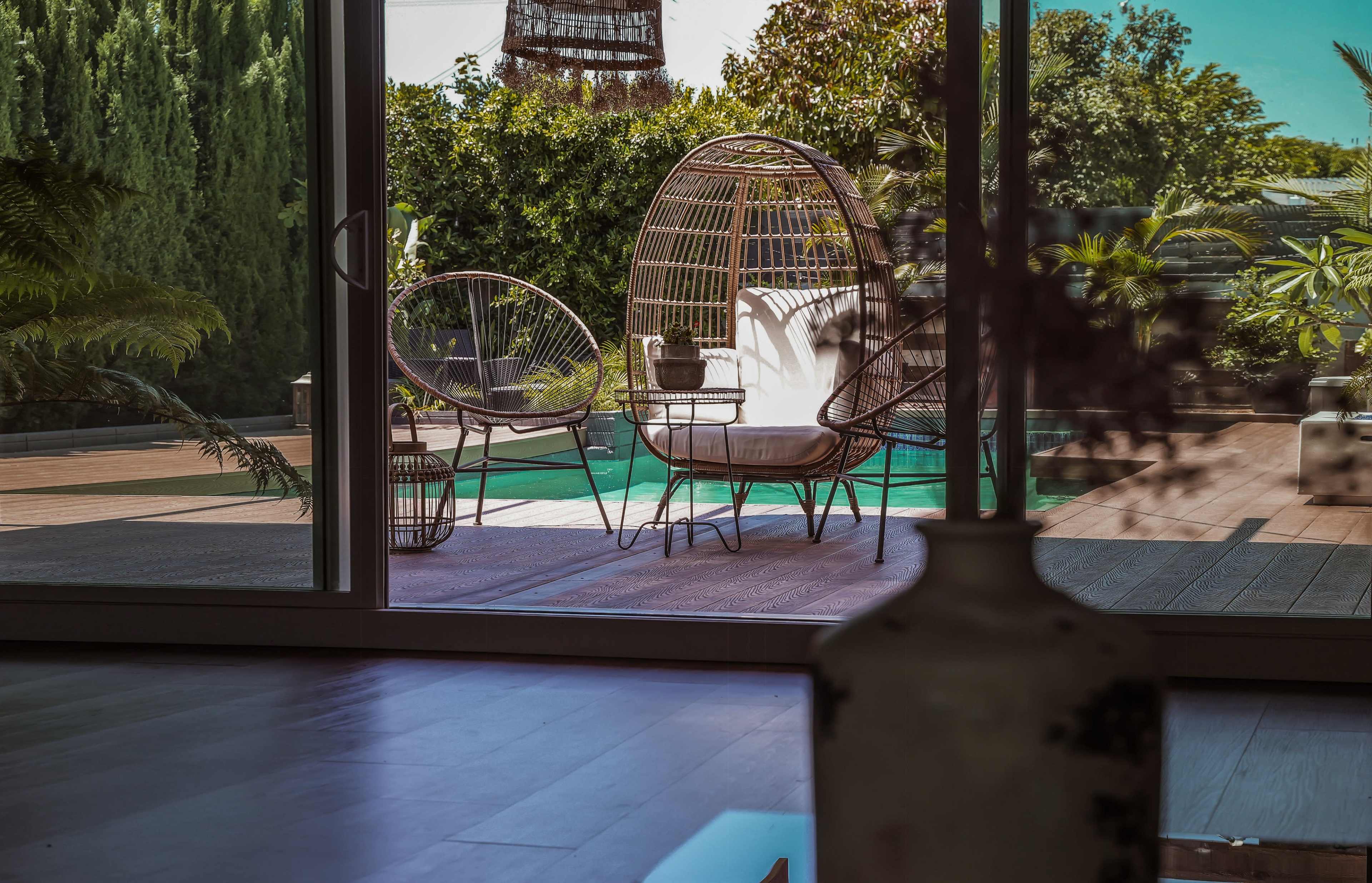 A cozy outdoor seating area is framed by large glass doors, featuring two woven chairs beside a pool surrounded by lush greenery.
