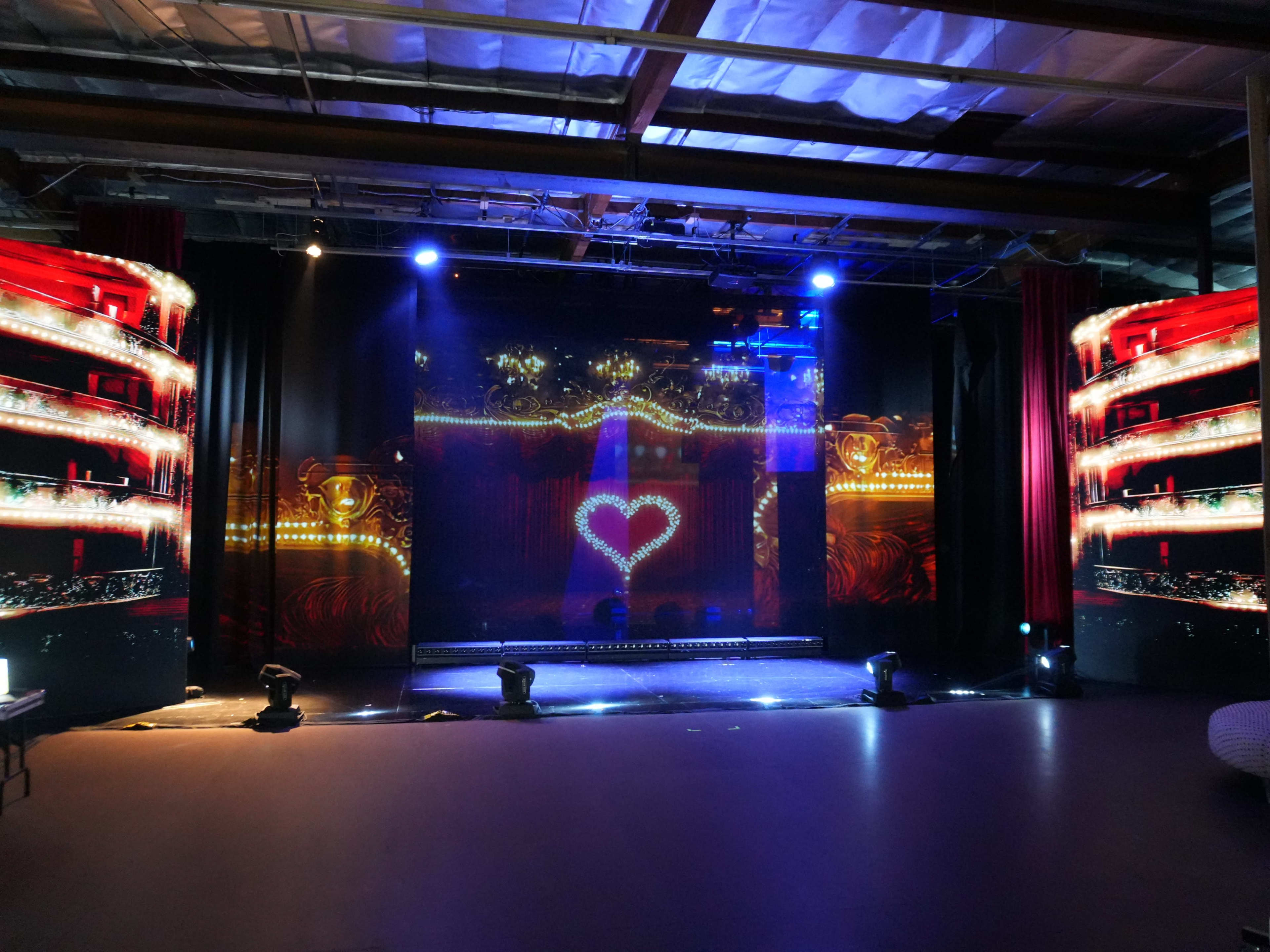 A brightly lit stage features large digital screens displaying colorful graphics, including a heart shape, surrounded by theatrical lighting.