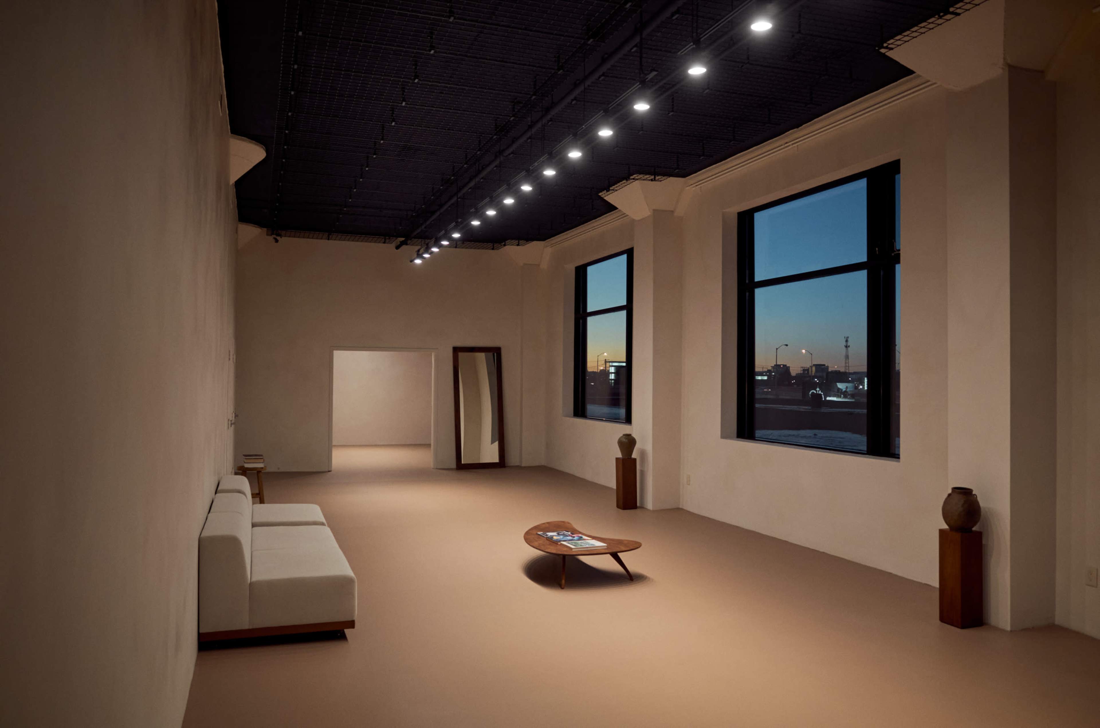 A spacious, minimalistic room features a white sofa, a low wooden coffee table, and large windows showcasing a sunset view.