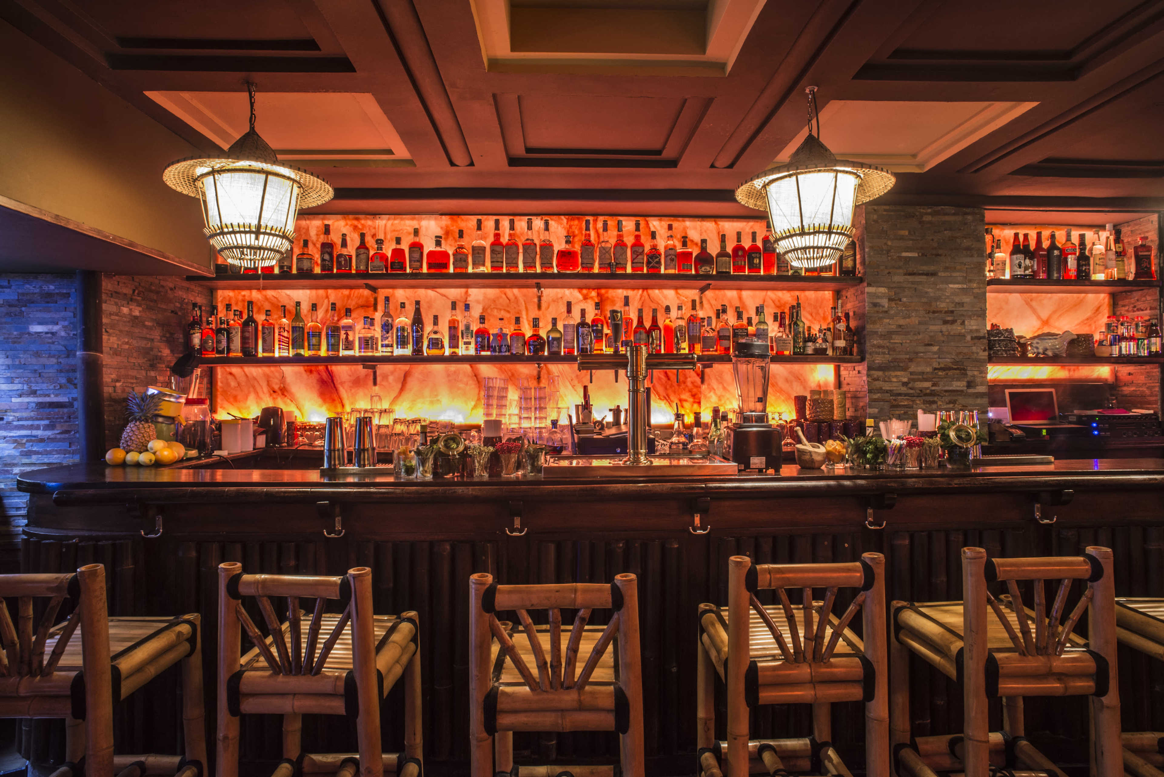 The image shows a wooden bar with several high stools, illuminated by two pendant lights and lined with shelves filled with various bottles of liquor.