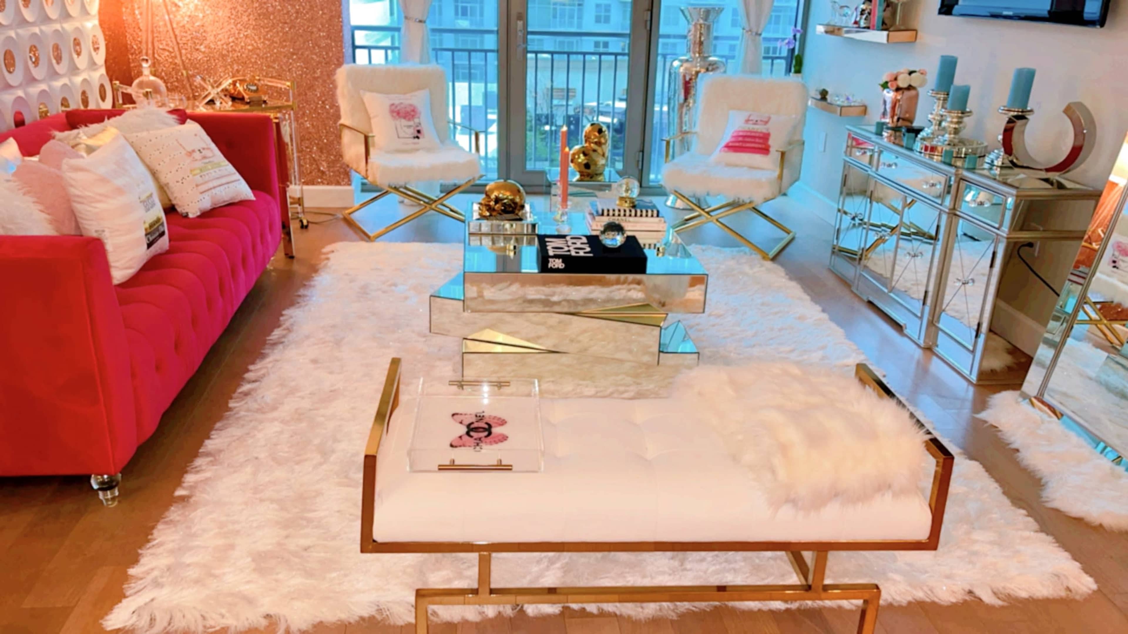The room features a stylish living area with a pink sofa, gold-accented chairs, a mirrored coffee table, and a white shag rug.