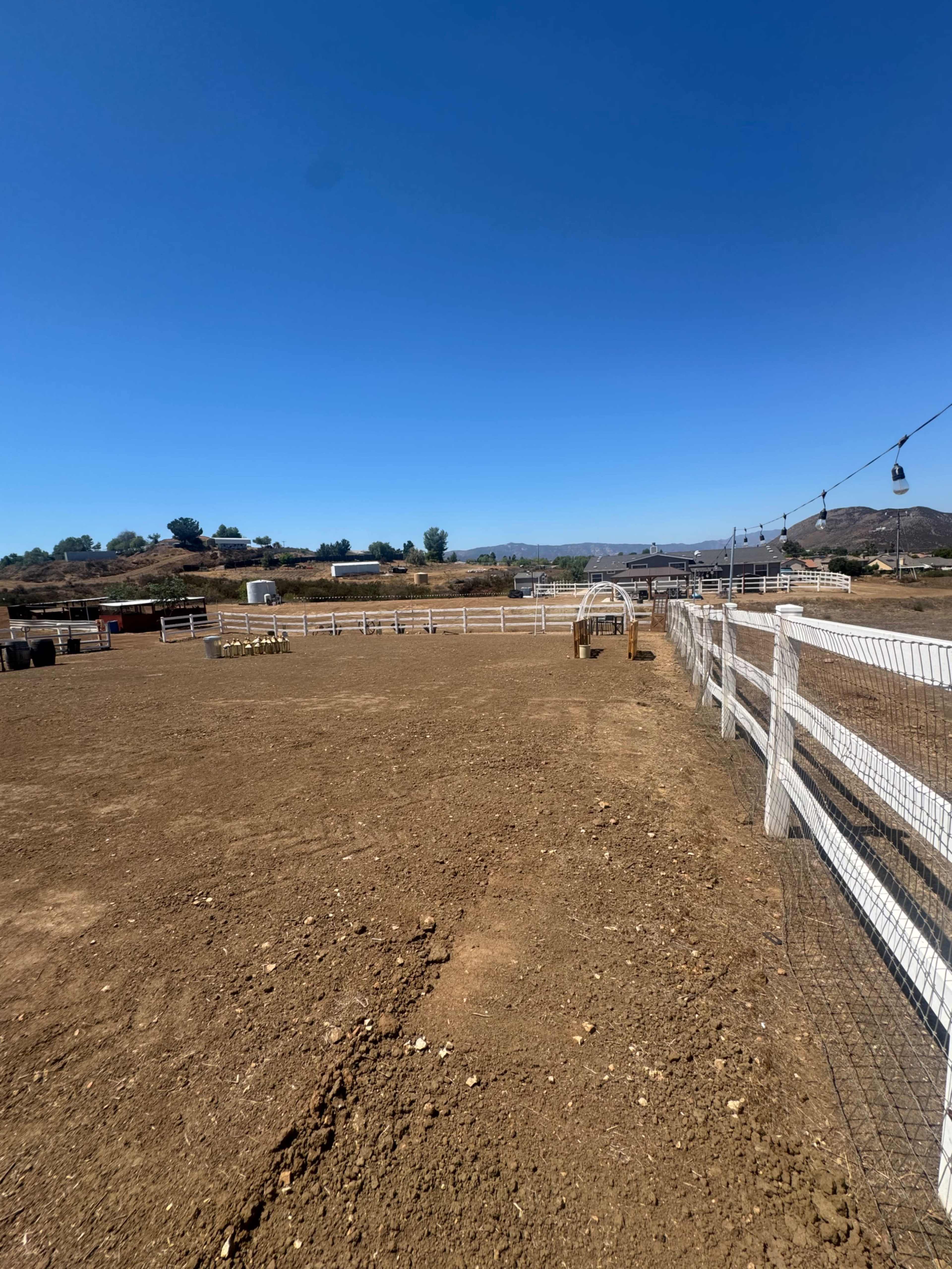 The image shows an expansive dirt horse arena with a white fence and distant hills under a clear blue sky.