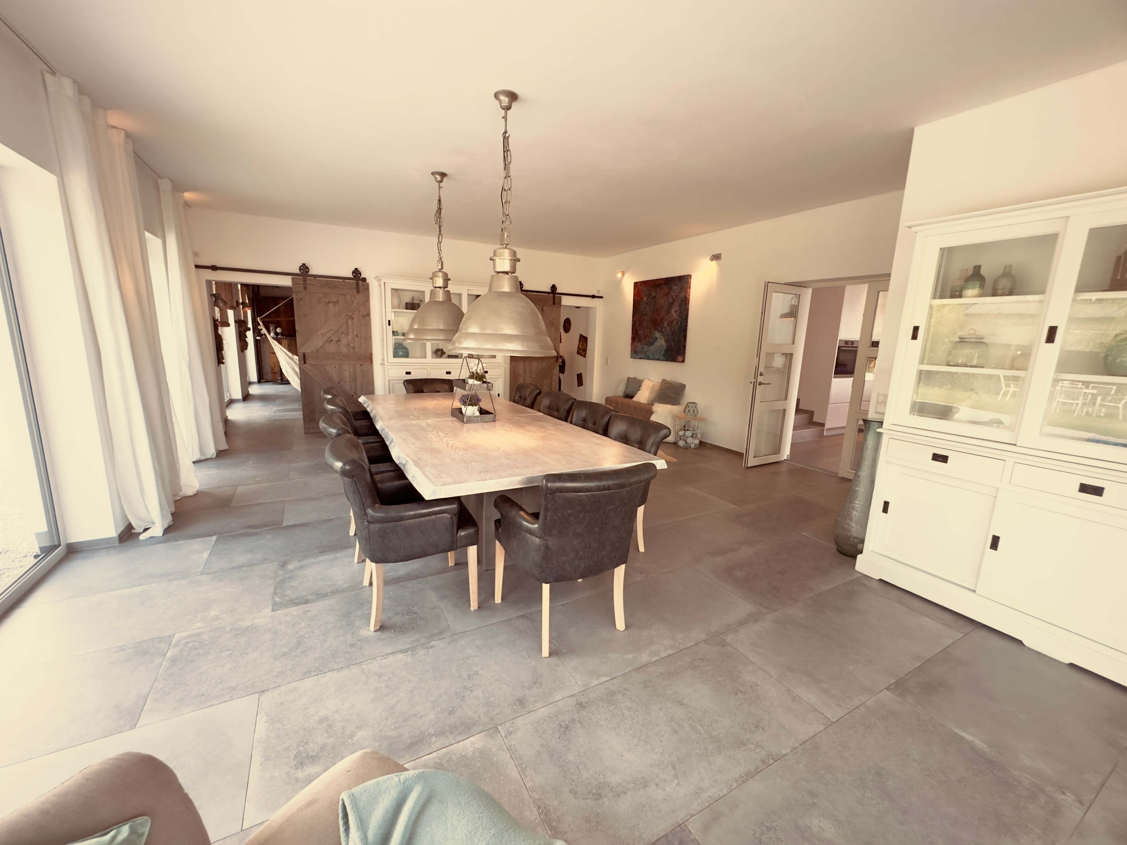 The image shows a spacious indoor dining area with a long wooden table and leather chairs, featuring large windows that allow natural light to fill the room.