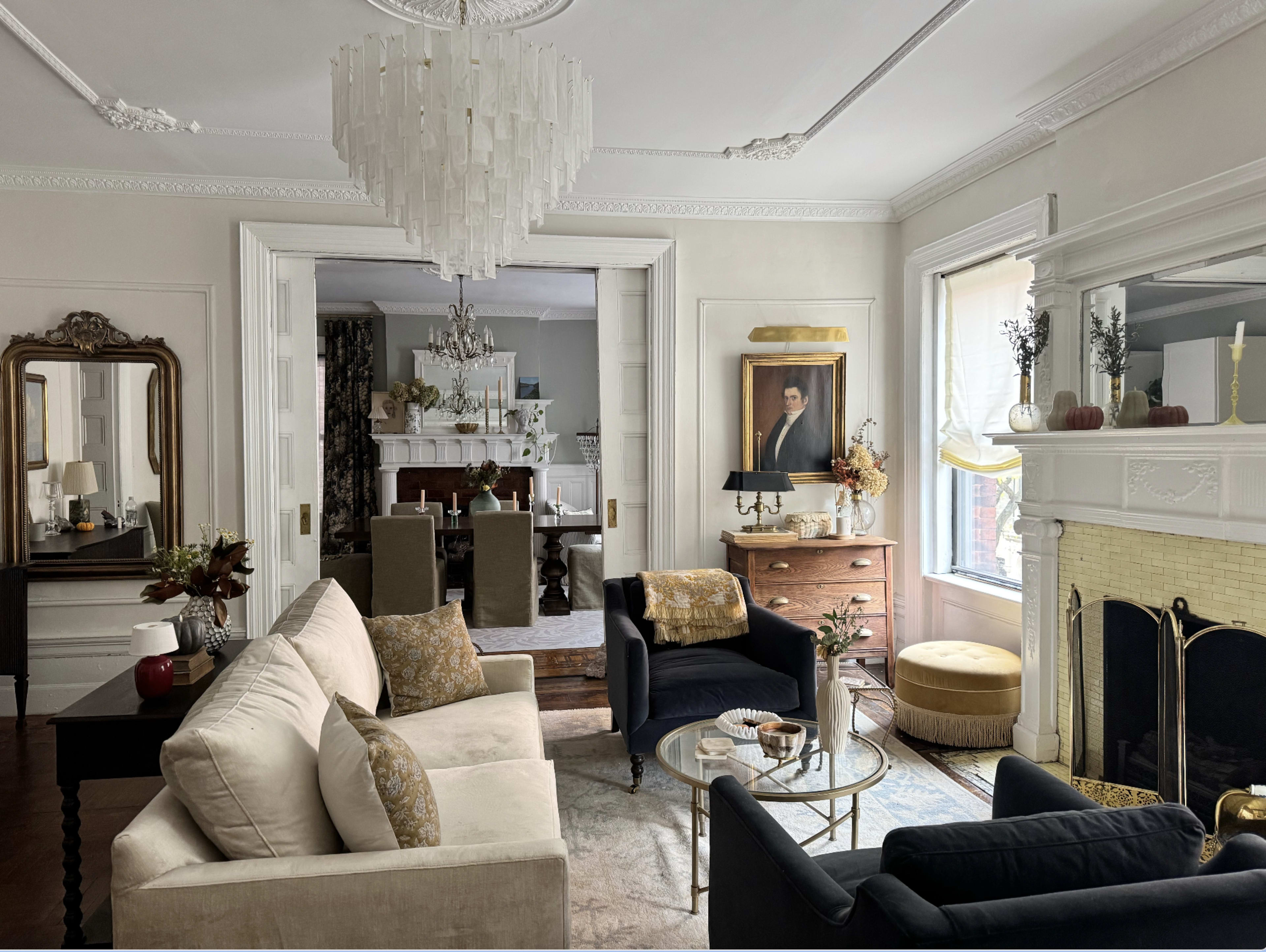 A living room features a chandelier, a fireplace with a yellow drapery, a mirror, and a portrait on the wall, with a mix of modern and vintage furniture.