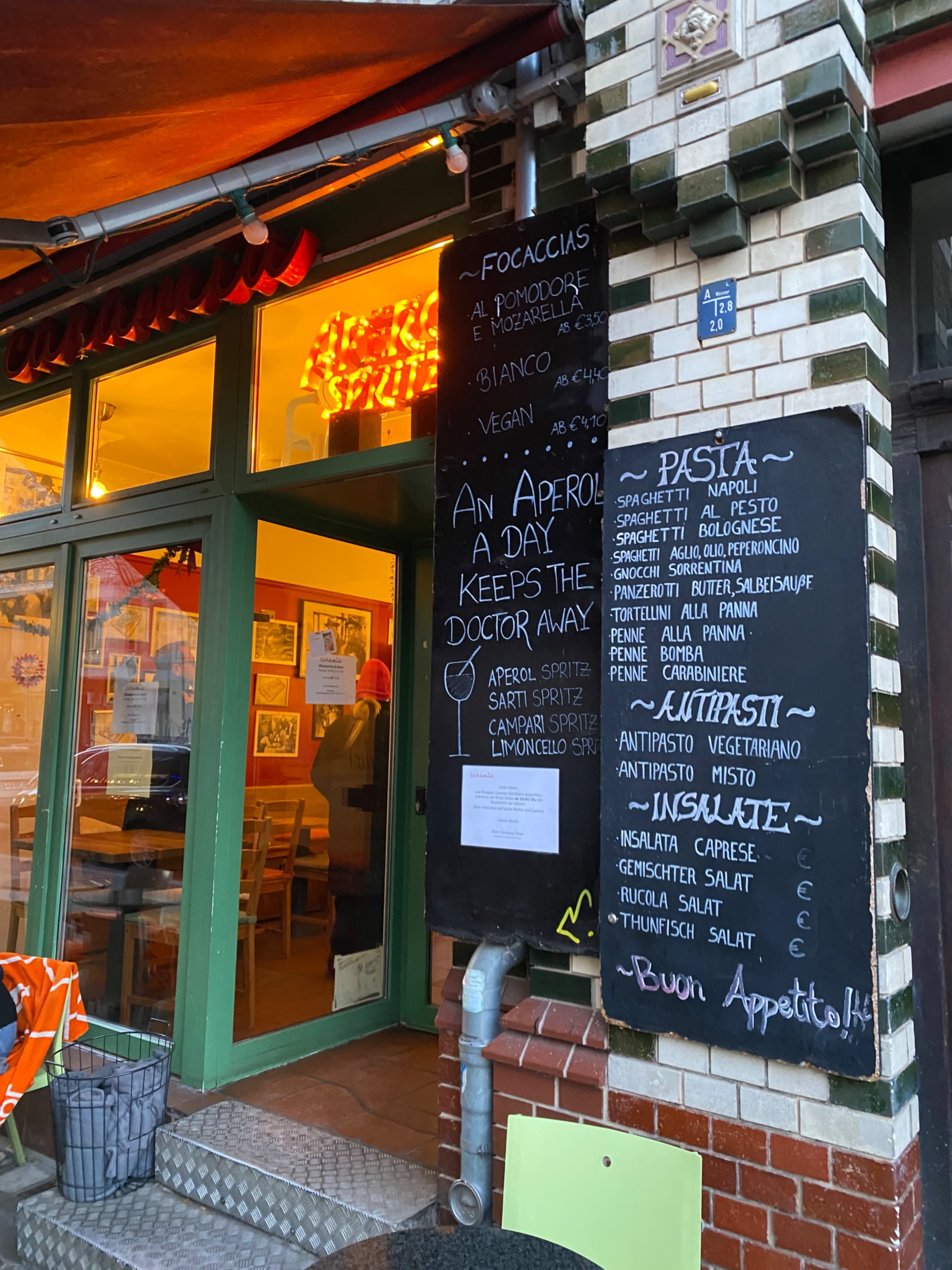 The image shows a restaurant entrance with a menu board featuring various Italian dishes written in chalk on a wall.