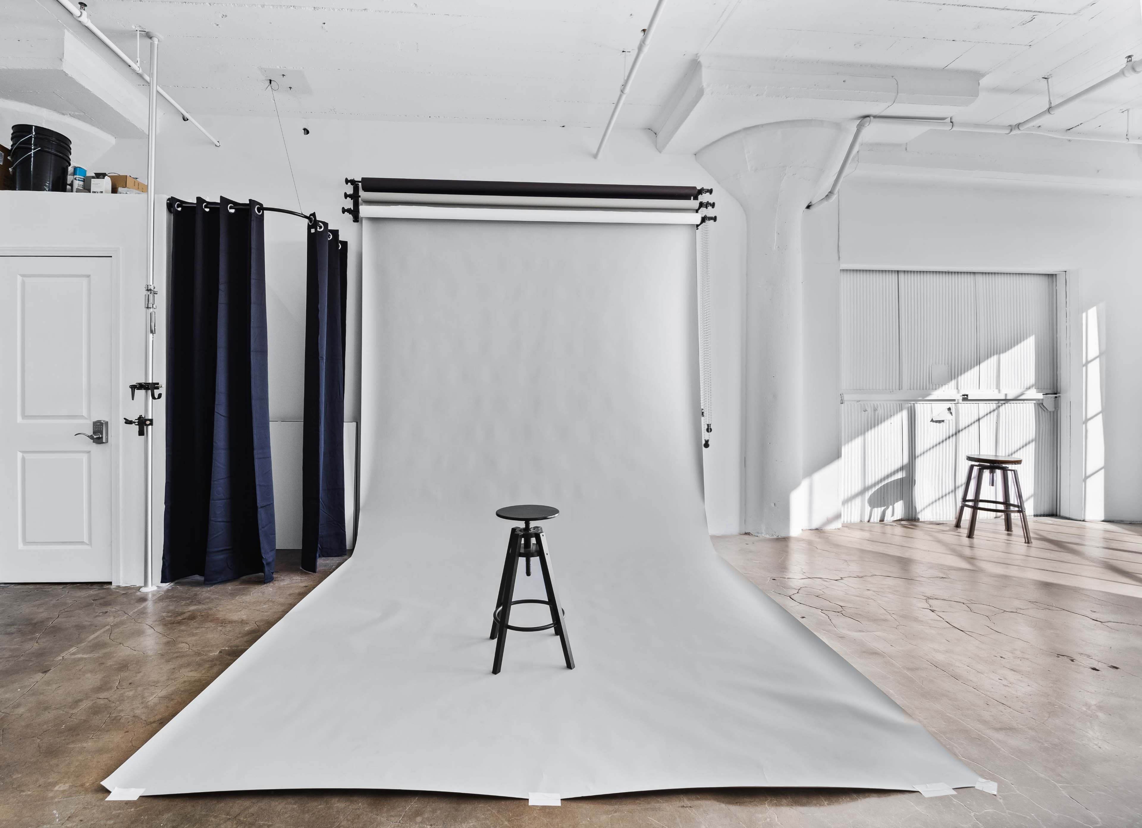 A simple photography studio is set up with a gray backdrop, a black stool in the center, and natural light streaming through large windows.