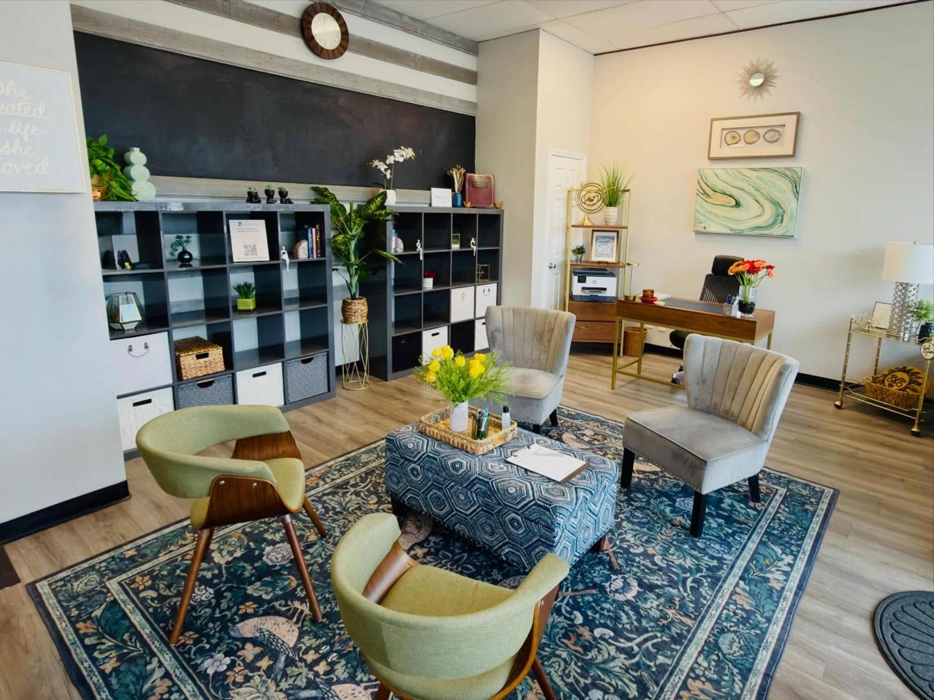 The image shows a modern office lounge area with four chairs, a decorative rug, a bookshelf, and a wooden desk decorated with plants and artwork.