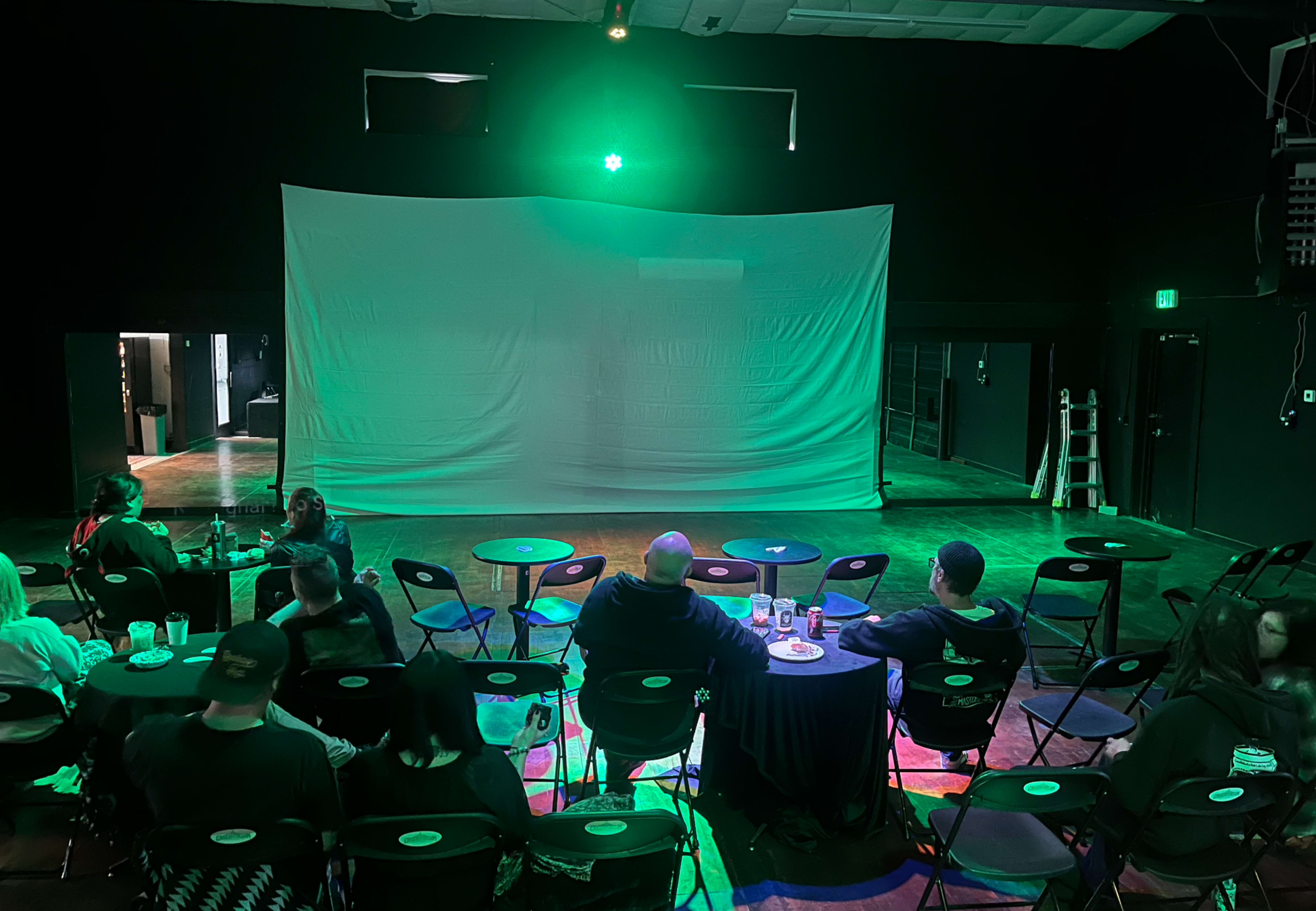 A small audience sits at tables facing a large blank screen illuminated by green stage lighting in a darkened performance space.