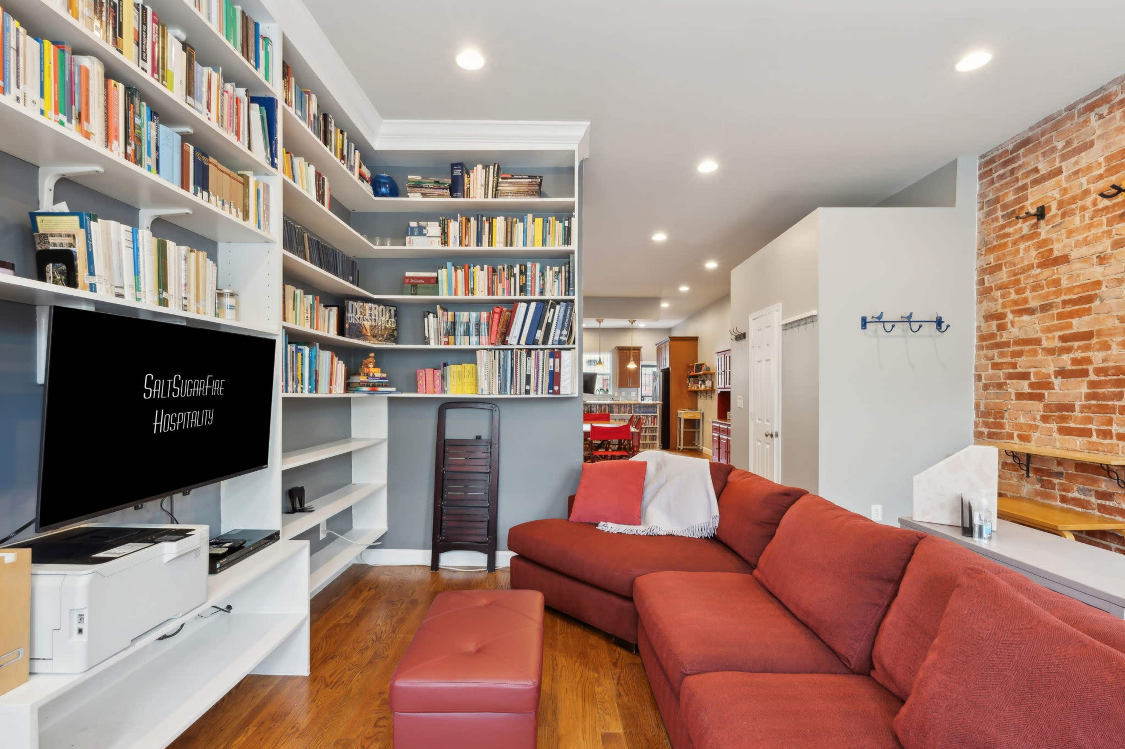 A red sectional couch faces a television on a shelf, with a tall bookcase filled with books and a brick wall in the background.