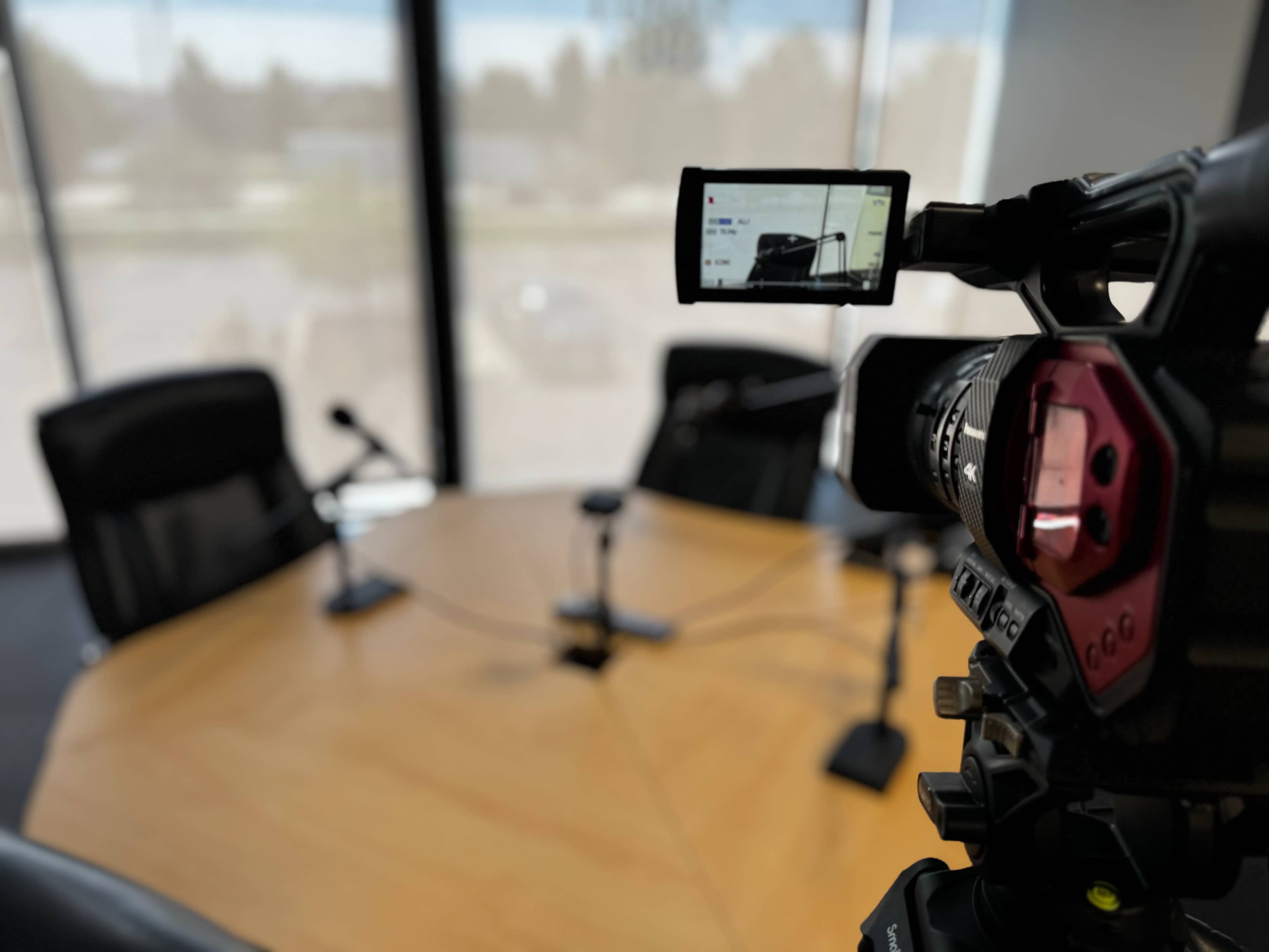 A video camera is positioned on a tripod facing a large wooden conference table with two microphones set up, inside a room with large windows.