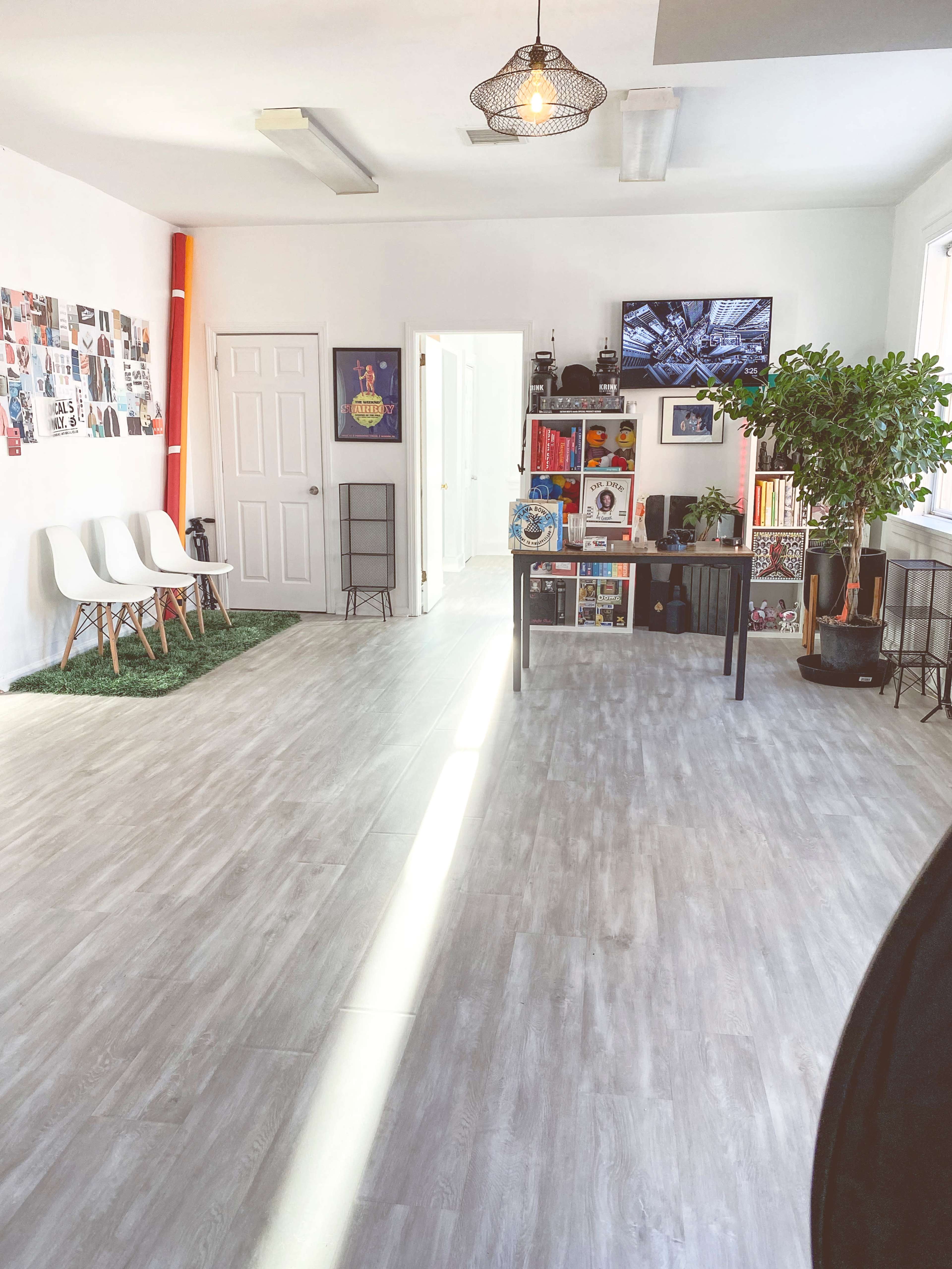 A bright, modern workspace features three white chairs, a table with decorative items, and a large wall-mounted television, with vibrant artwork and plants throughout.