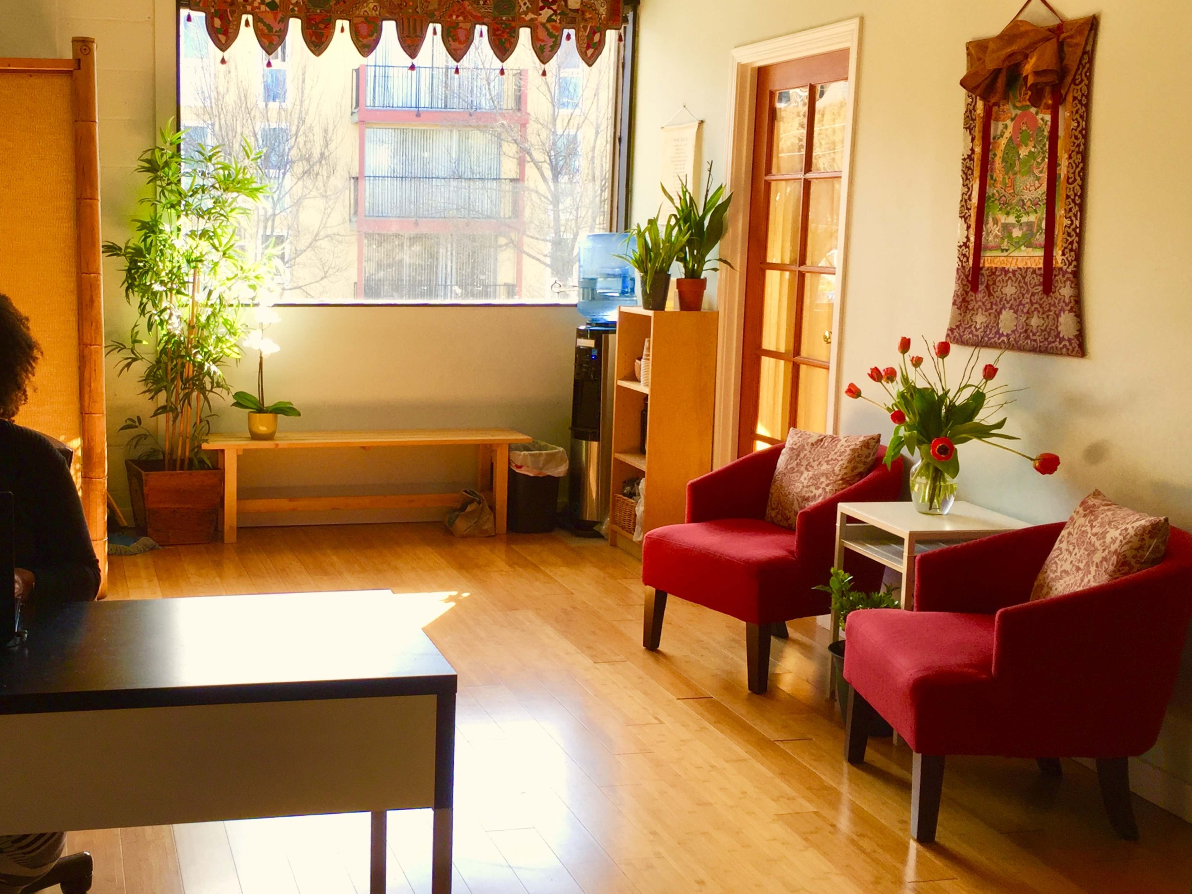 A bright room with wooden floors, a water cooler, two red chairs with decorative cushions, and a small table beside a flower arrangement.