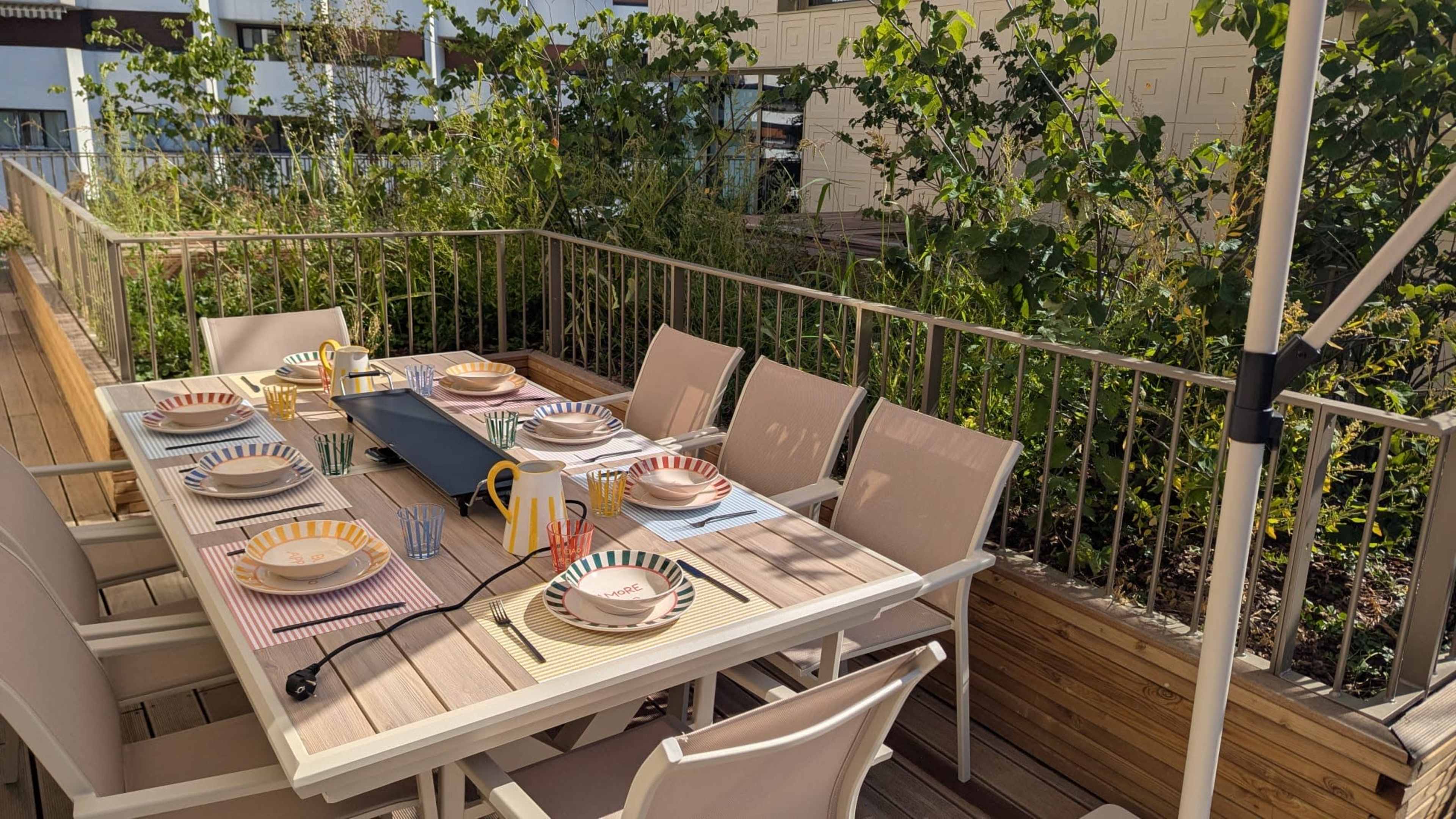A dining table is set with plates and utensils on a wooden deck surrounded by greenery.