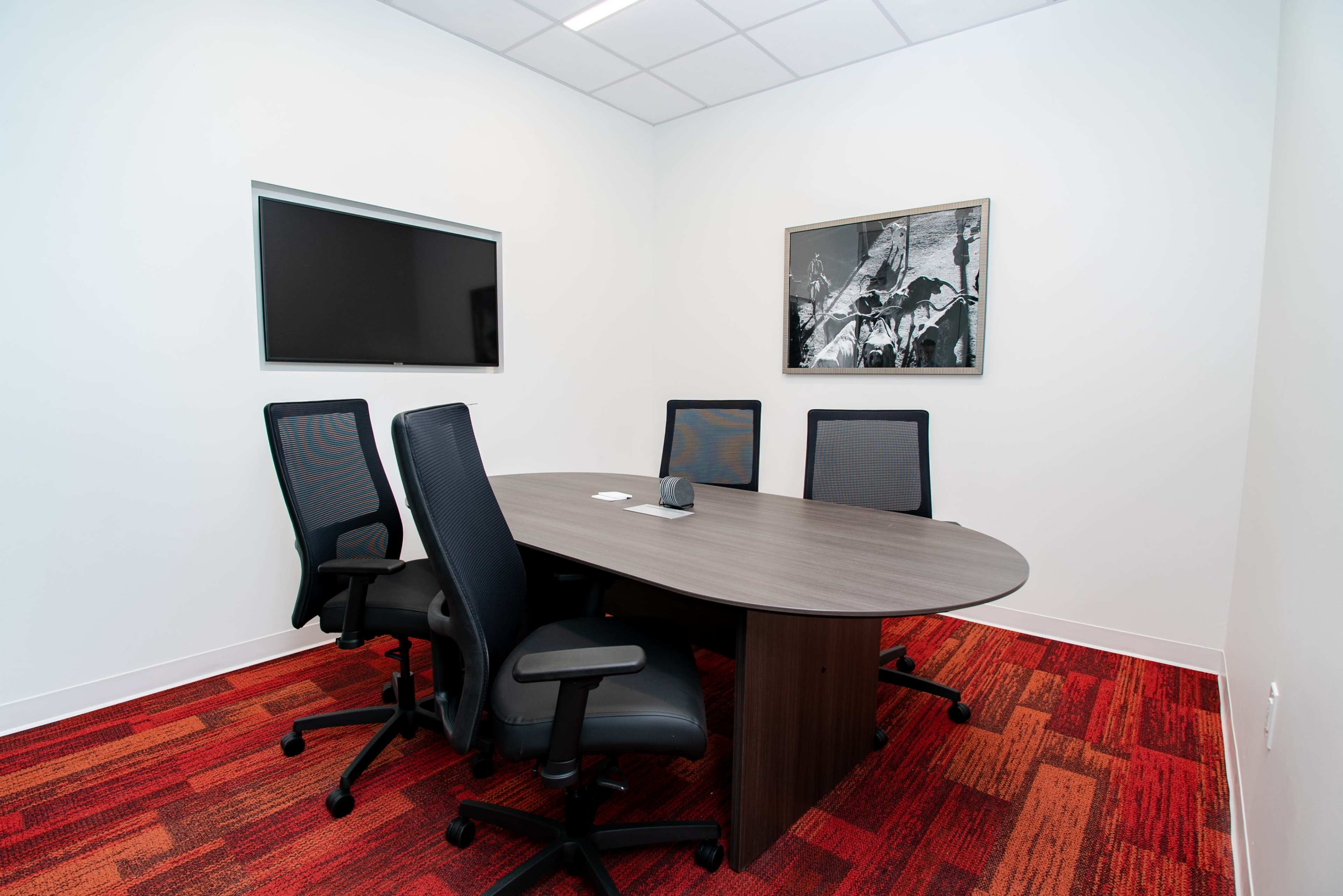 A small conference room with a round table, four black office chairs, a mounted television, and a wall art piece against a white background.