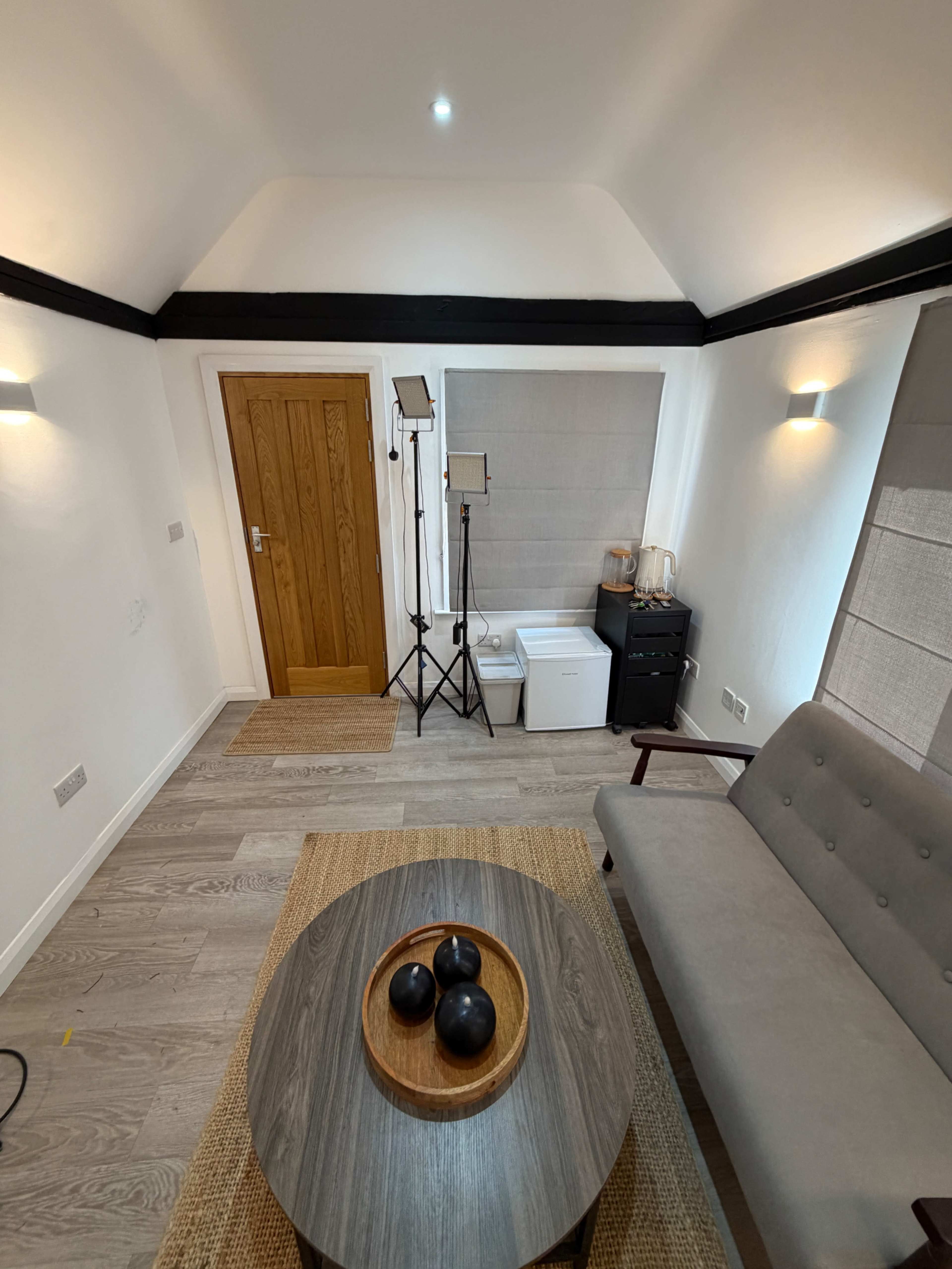 The image shows a minimalist interior room featuring a gray couch, a wooden coffee table with three black decorative balls, a window with shades, and a standing light beside a door.