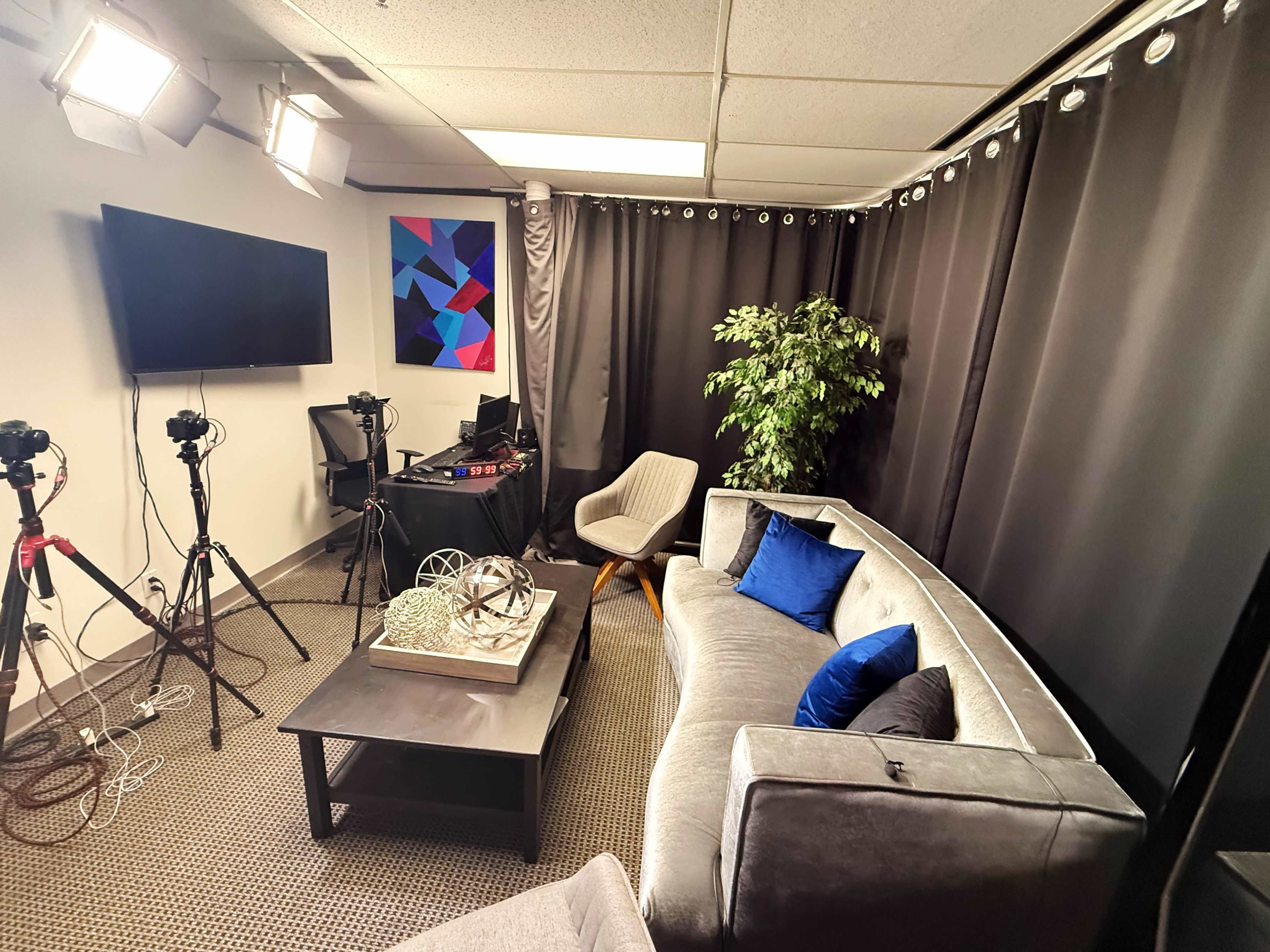 The image shows a cozy studio setup featuring a gray couch, a coffee table with decorative items, a potted plant, and several cameras on tripods.