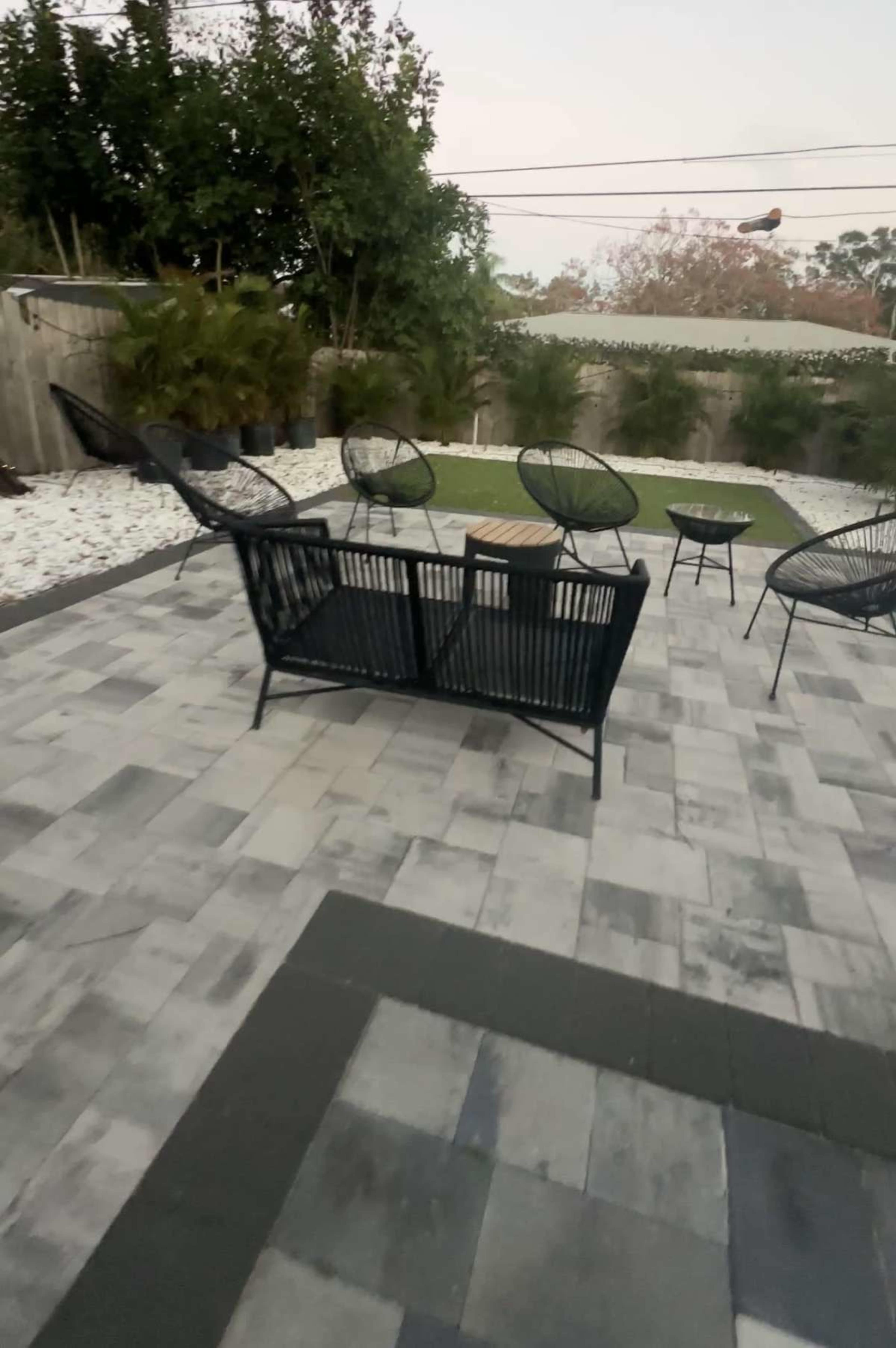 The image shows a patio area with black outdoor furniture, including a sofa and several chairs, surrounded by a landscaped garden and stone pathways.