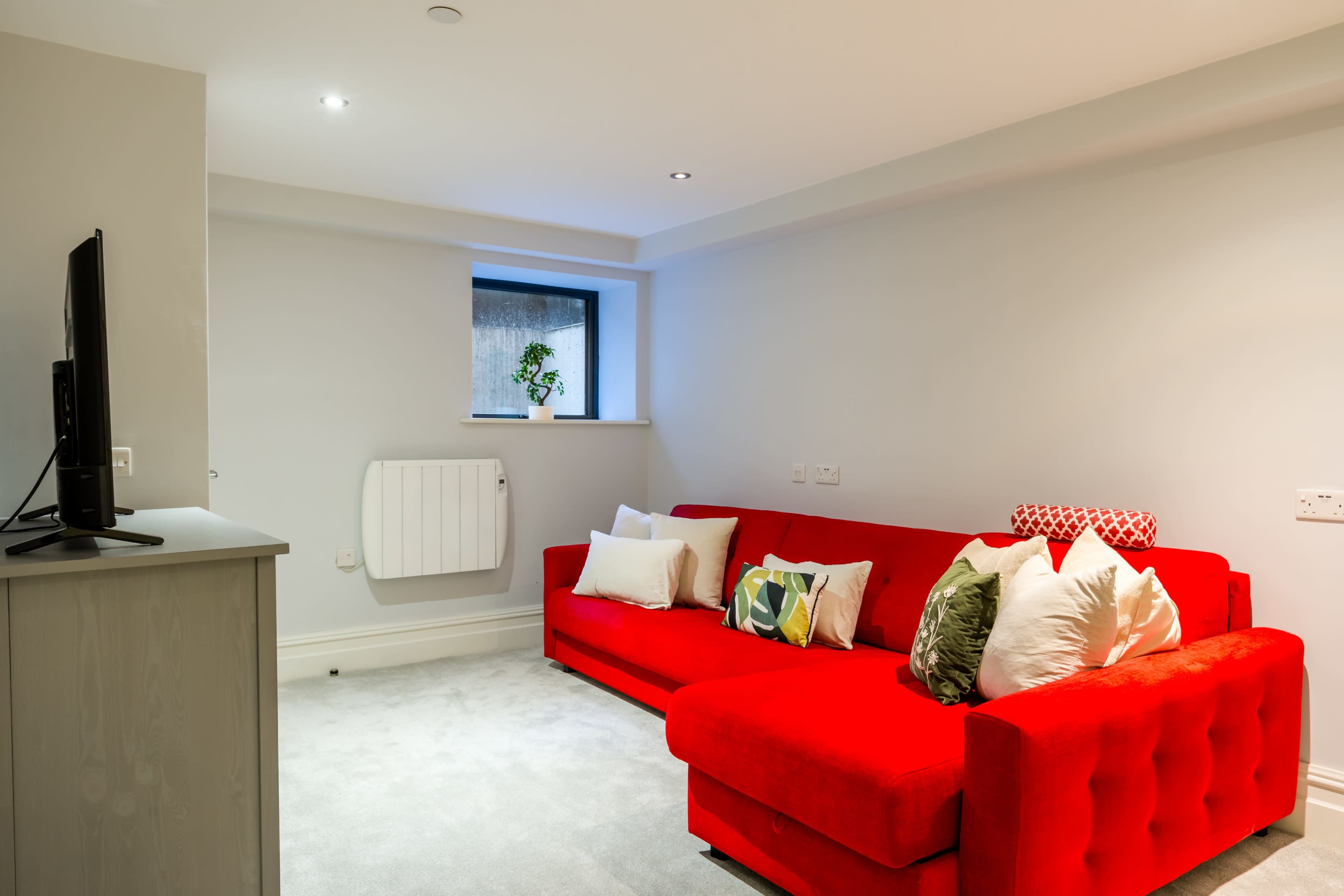 A red sectional sofa with decorative pillows is positioned against a light gray wall in a cozy living room.