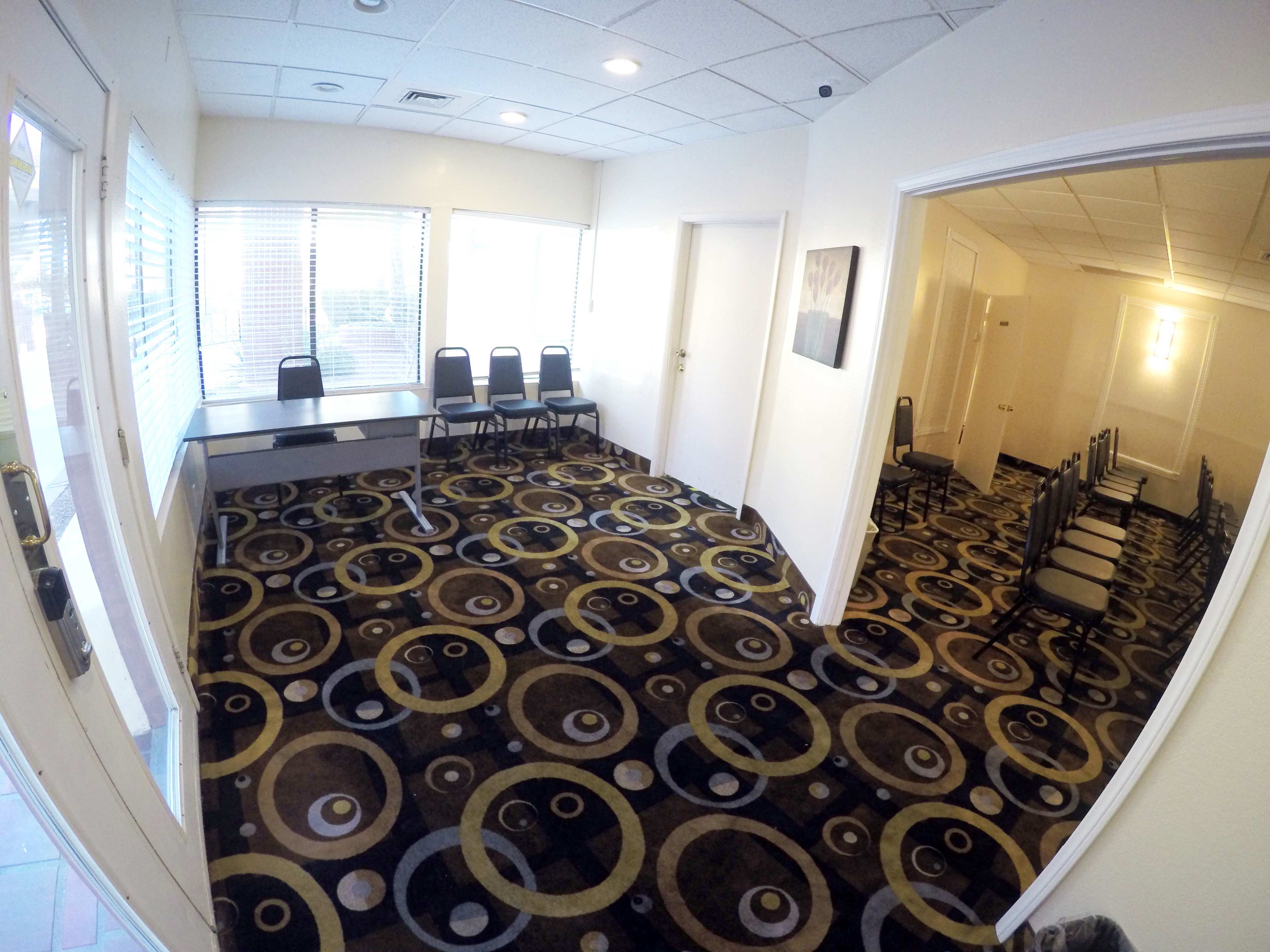 A spacious, well-lit meeting room featuring a circular-patterned carpet, a table at the front, and several rows of chairs arranged along the sides.