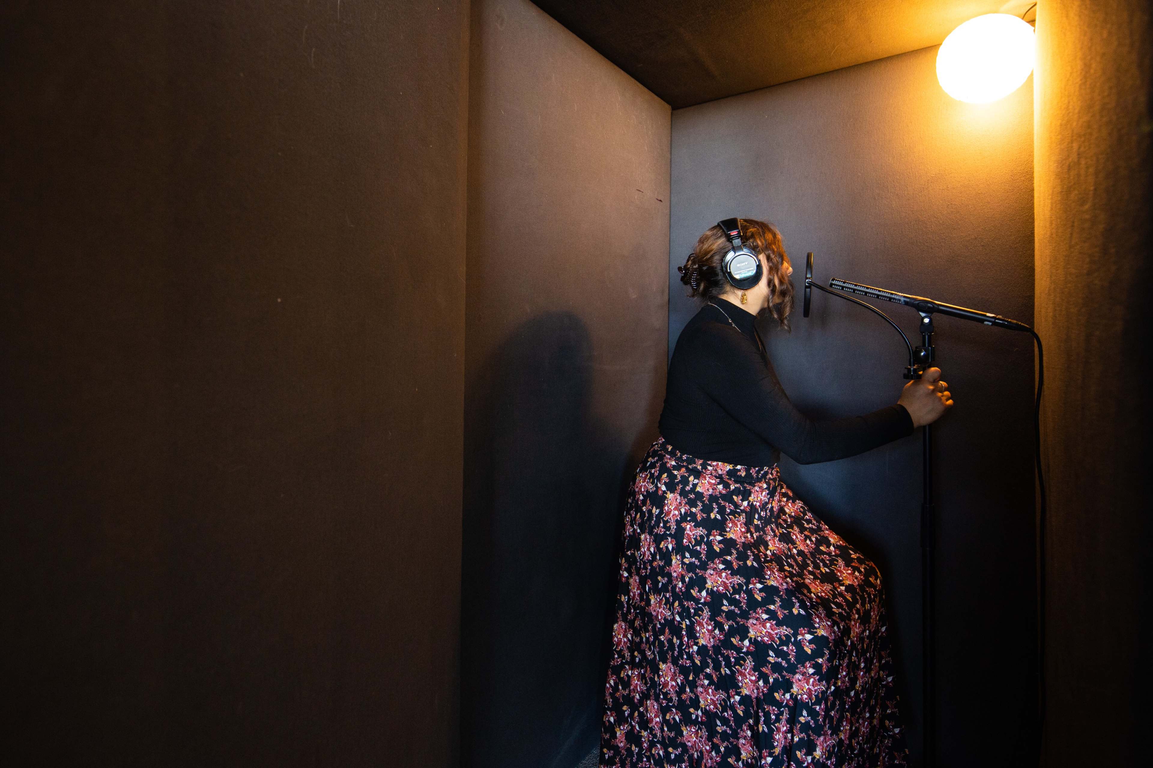 A person wearing headphones stands in a vocal booth while adjusting a microphone.