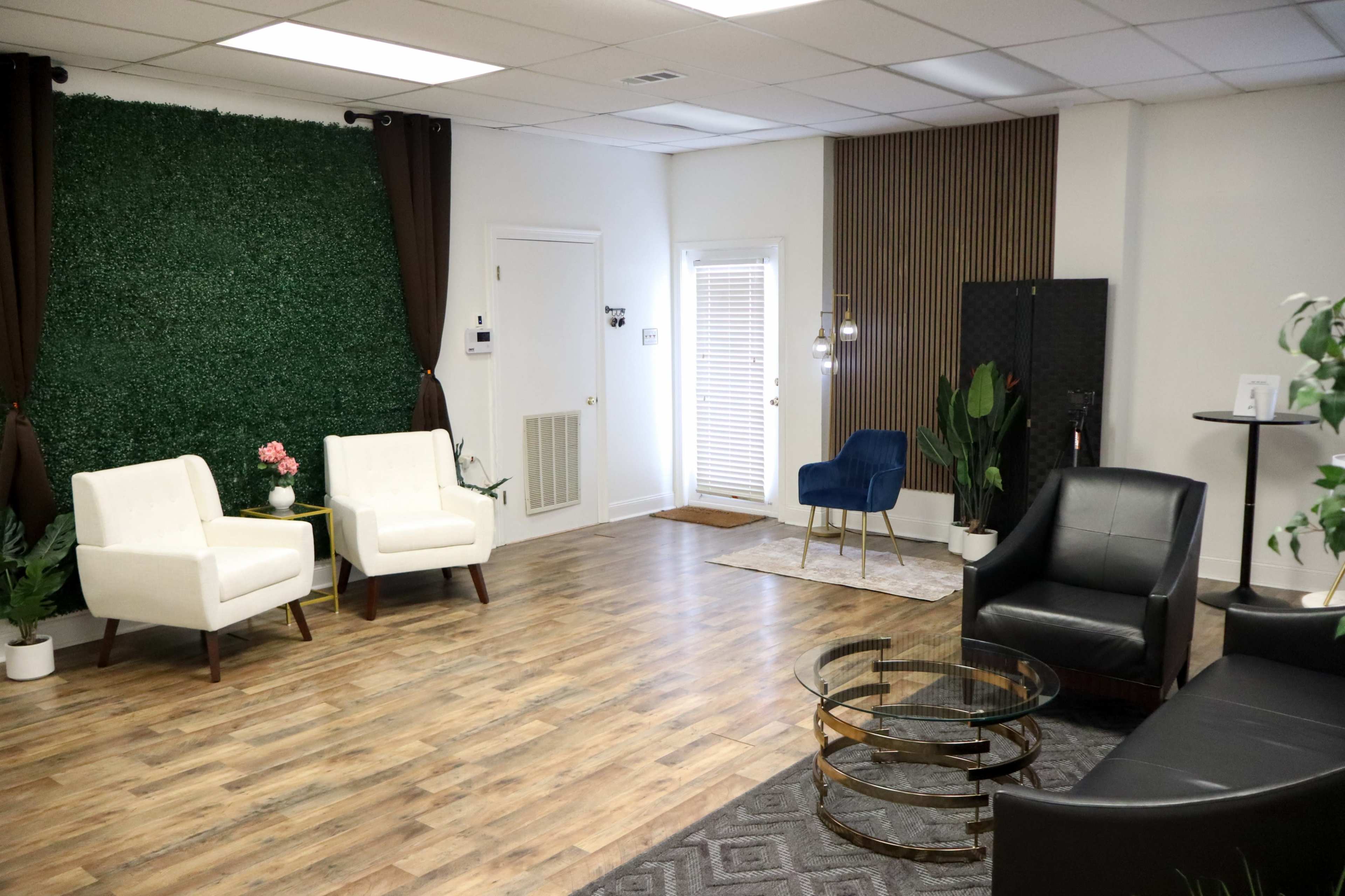 The image shows a spacious interior room with a mix of modern seating, greenery on one wall, and a variety of plants placed throughout.