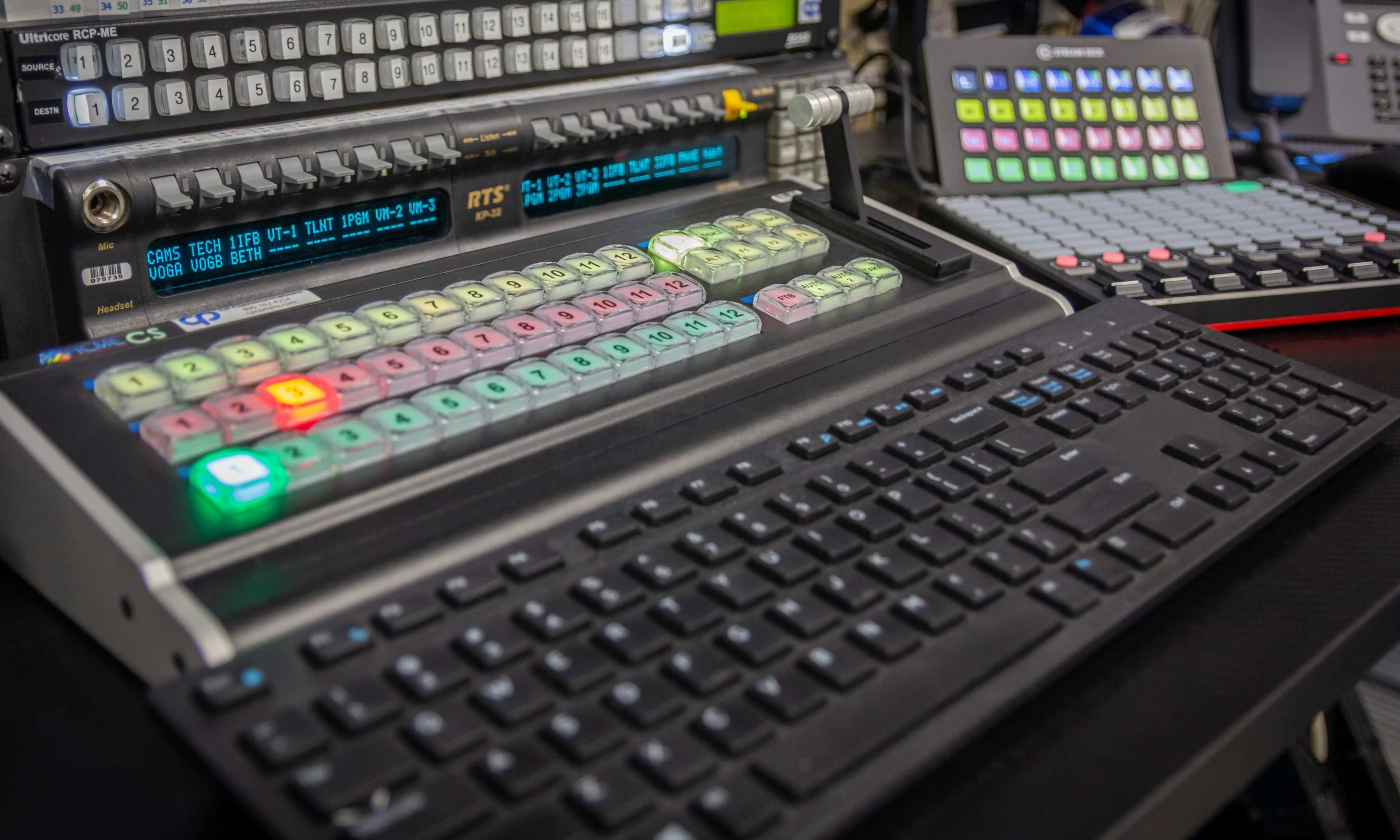 A high-tech broadcast control panel with illuminated buttons and keyboards arranged on a workspace.