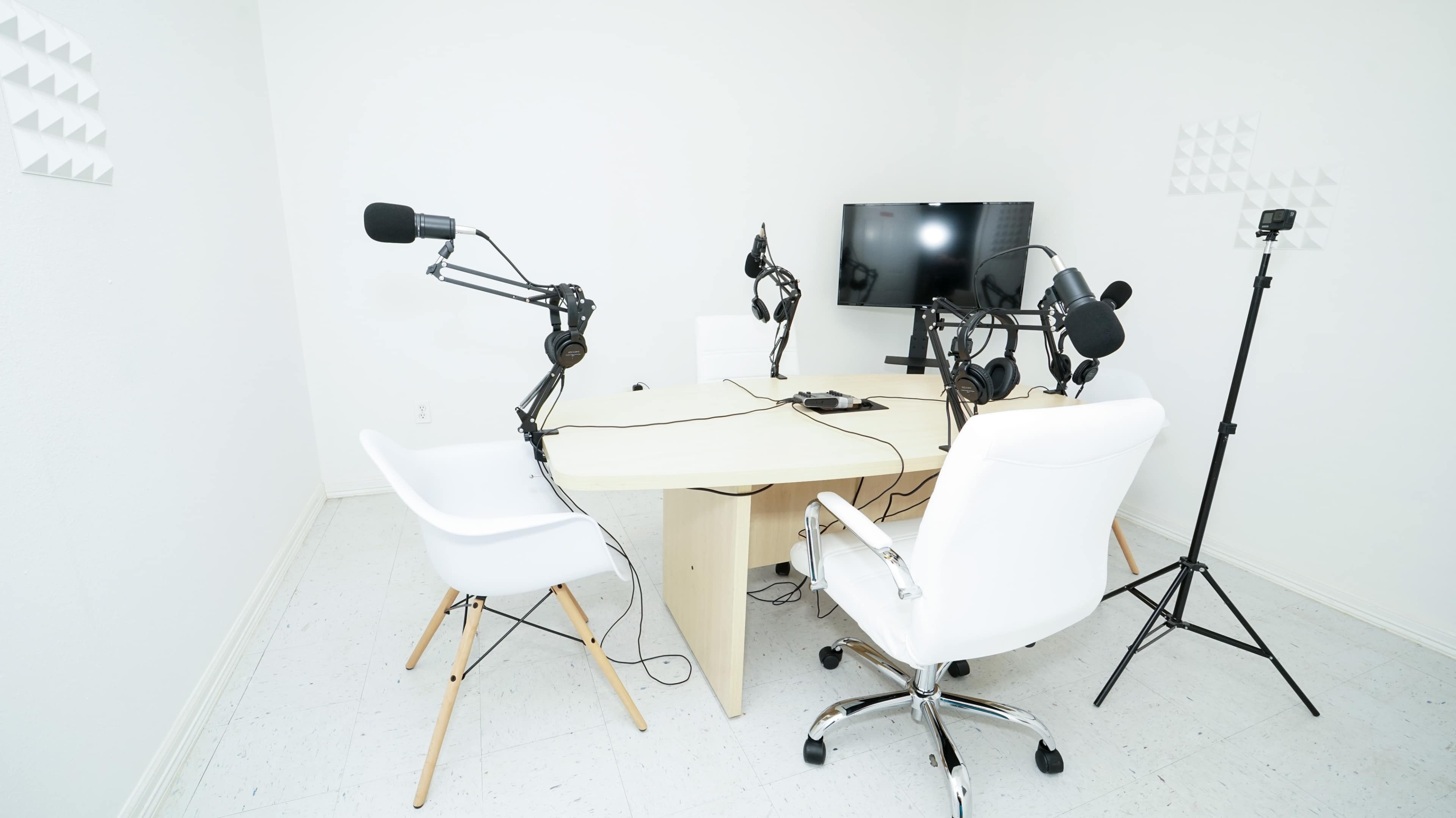 A minimalistic podcasting studio features a table surrounded by four white chairs, equipped with microphones mounted on adjustable arms and a television mounted on the wall.