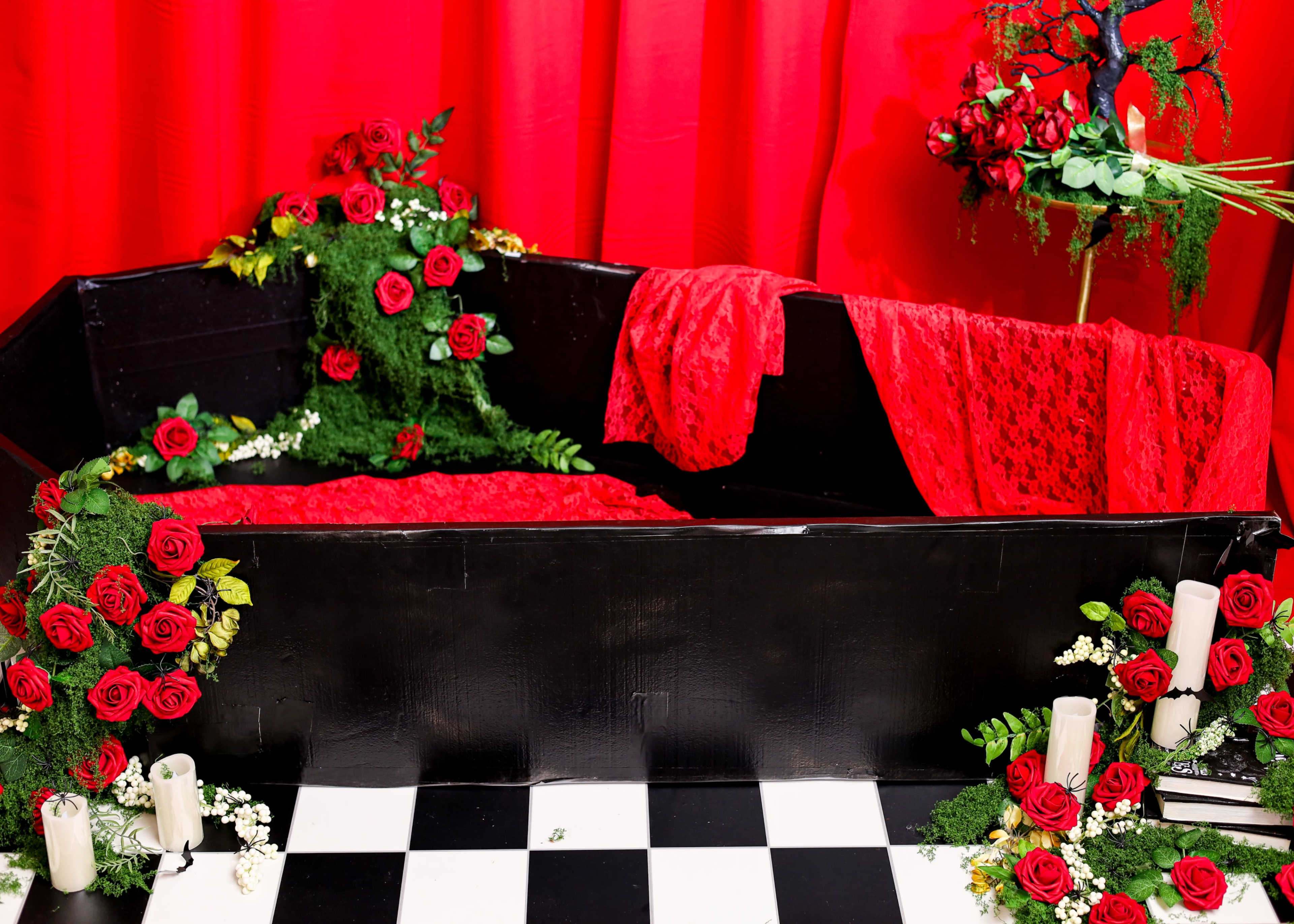 The image features a black coffin adorned with red roses and lace, set against a red backdrop, surrounded by candles and greenery.