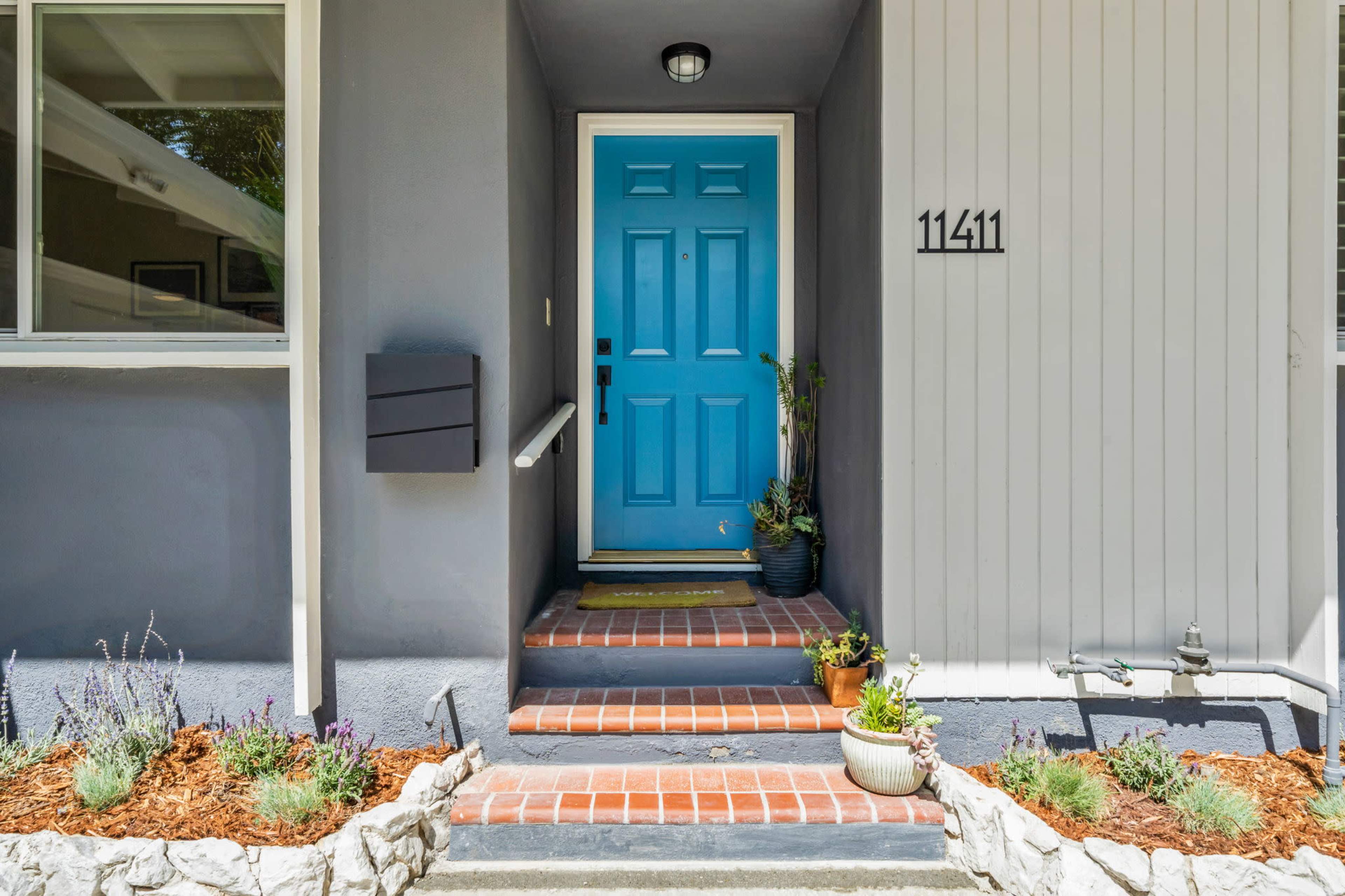 A front entrance with a bright blue door, brick steps, and potted plants on either side.